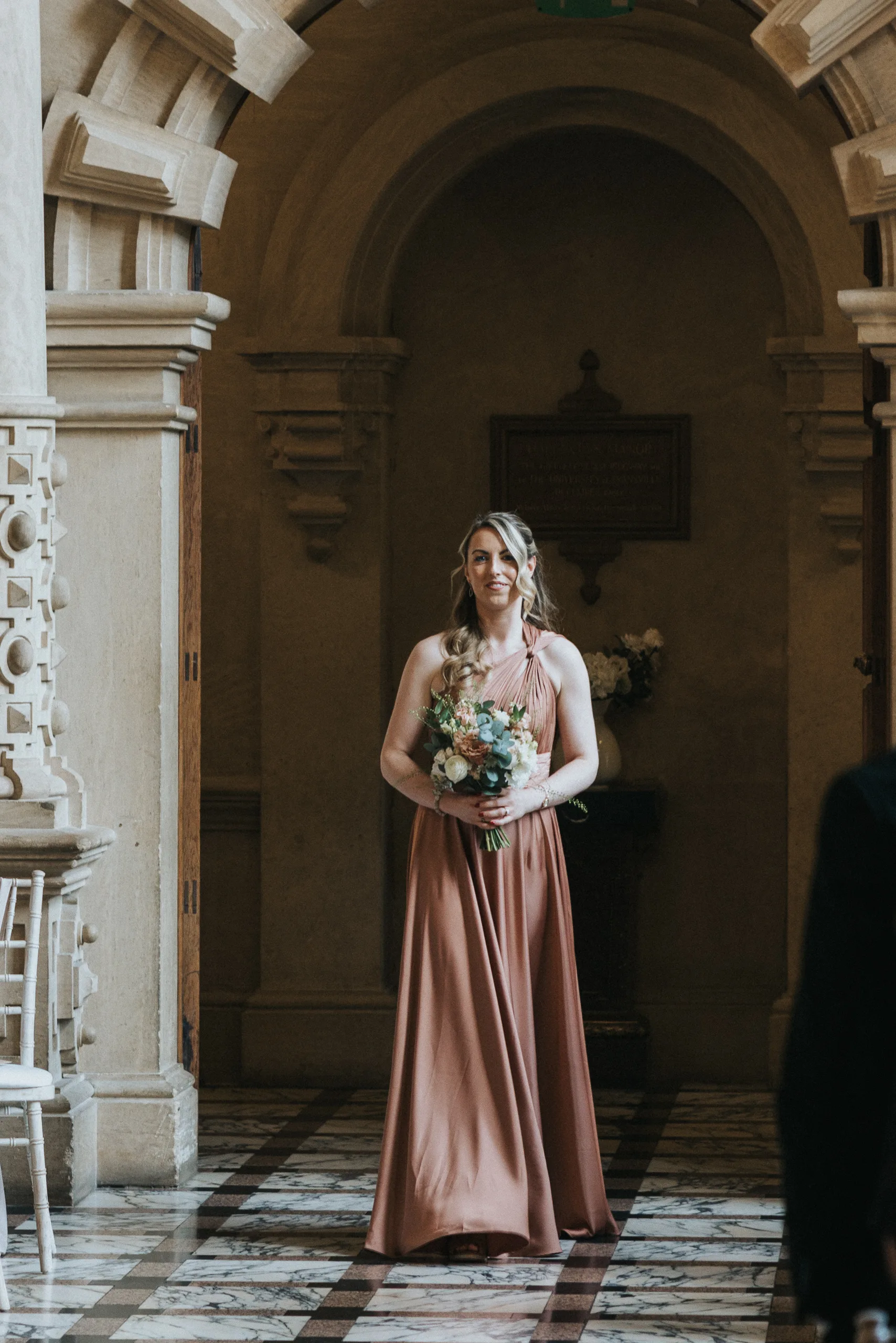 A woman with long blonde hair in loose curls, wearing a sleeveless rust-coloured gown, stands indoors holding a bouquet of flowers at a Harlaxton Manor wedding, framed by ornate stone arches, marble floors, and soft natural light. © Aimee Lince Photography, Lincolnshire photographer