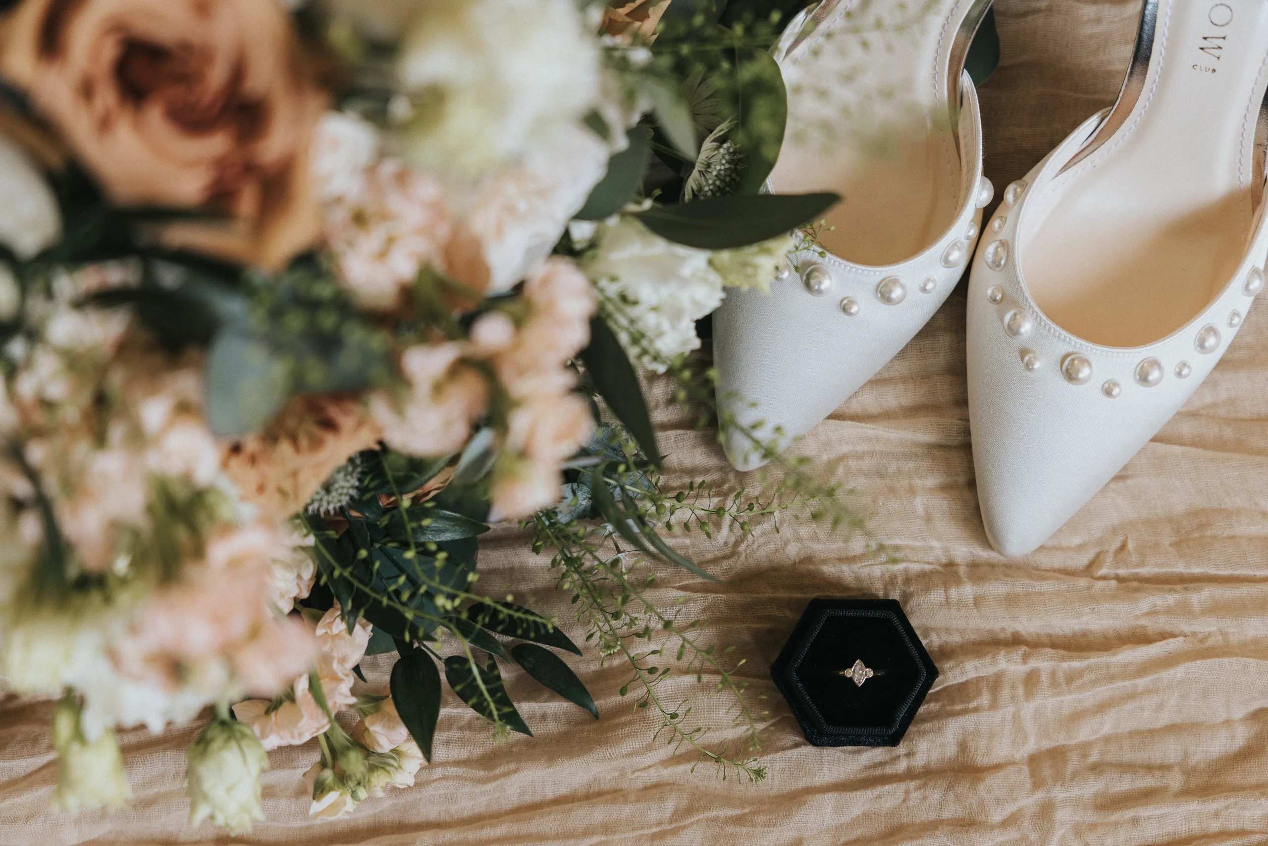 A close-up of white, pearl-adorned bridal shoes beside a bouquet of pastel flowers and greenery on beige fabric at a Harlaxton Manor Wedding. A black hexagonal ring box with a diamond ring sits near the shoes and bouquet. © Aimee Lince Photography, Lincolnshire photographer