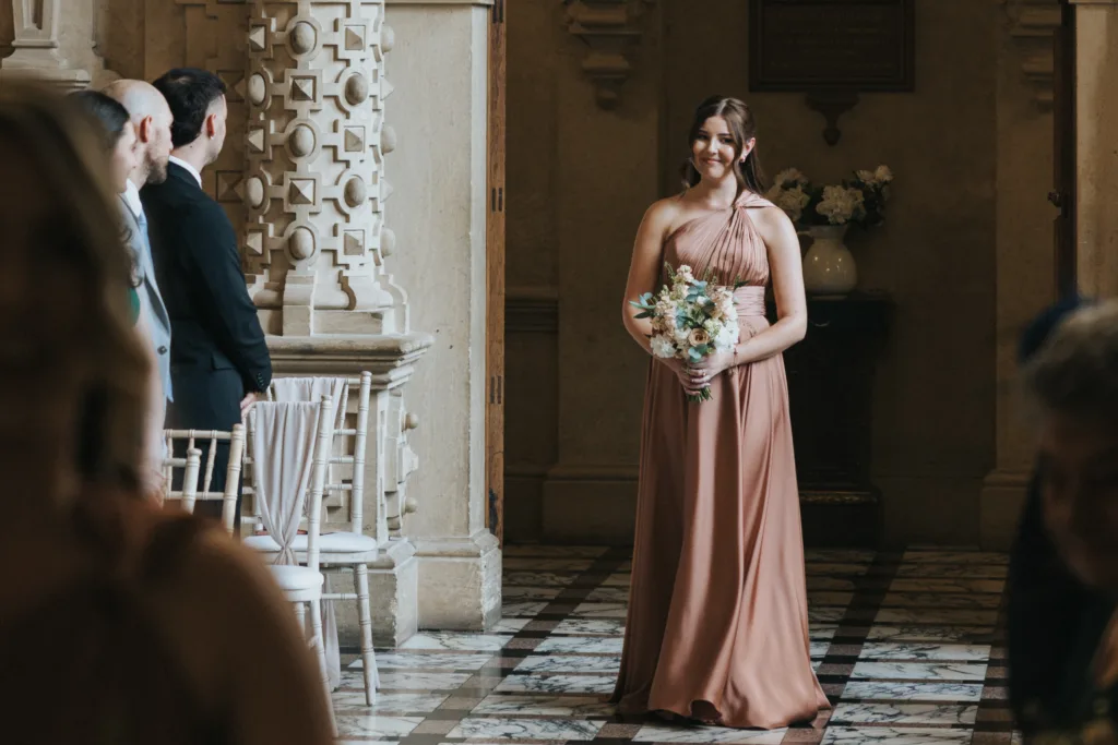 A woman in a rose-pink, one-shoulder gown holds a bouquet of flowers as she walks down the aisle at a Harlaxton Manor wedding, surrounded by marble floors, ornate columns, floral arrangements, and guests watching in the sunlit hall. © Aimee Lince Photography, Lincolnshire photographer