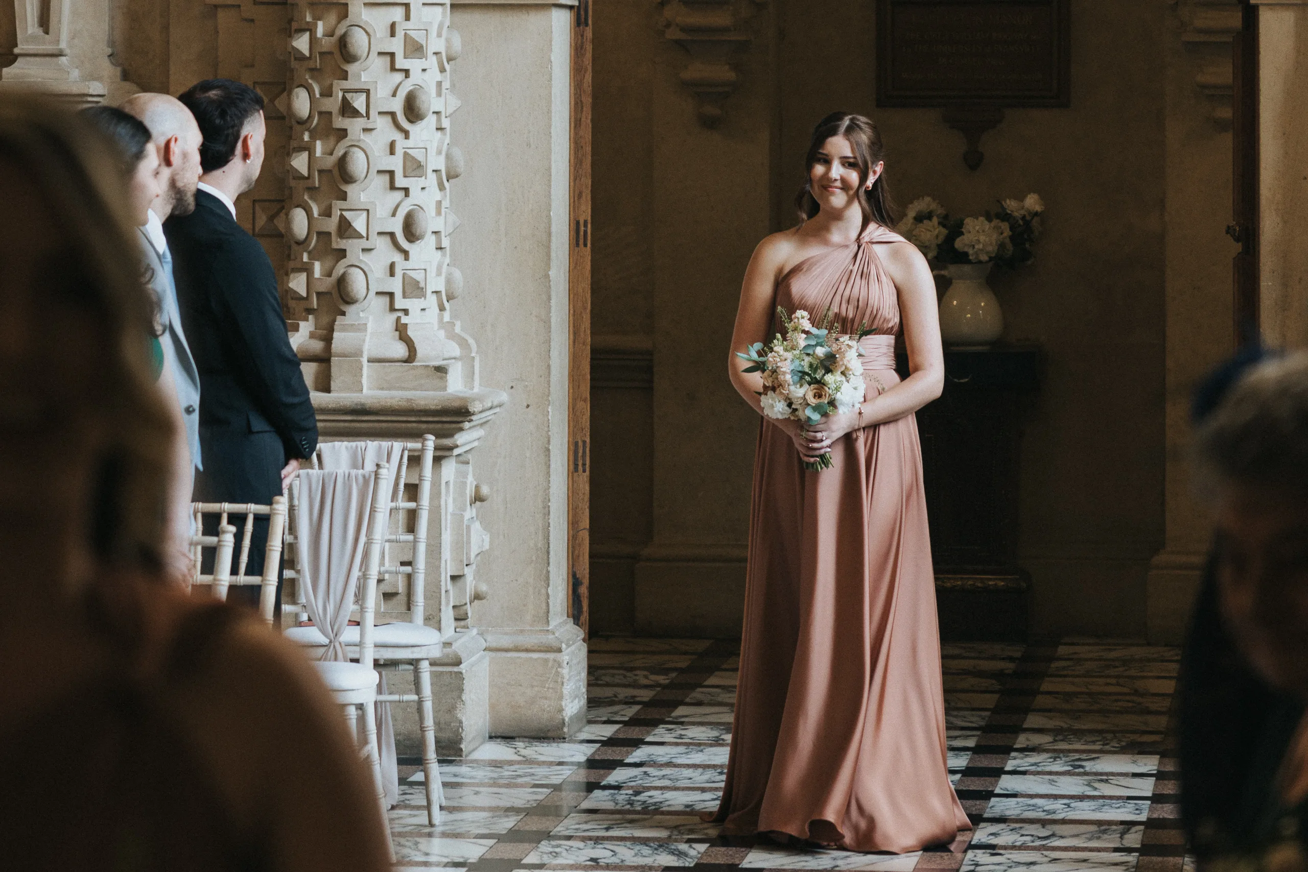A woman in a rose-pink, one-shoulder gown holds a bouquet of flowers as she walks down the aisle at a Harlaxton Manor wedding, surrounded by marble floors, ornate columns, floral arrangements, and guests watching in the sunlit hall. © Aimee Lince Photography, Lincolnshire photographer