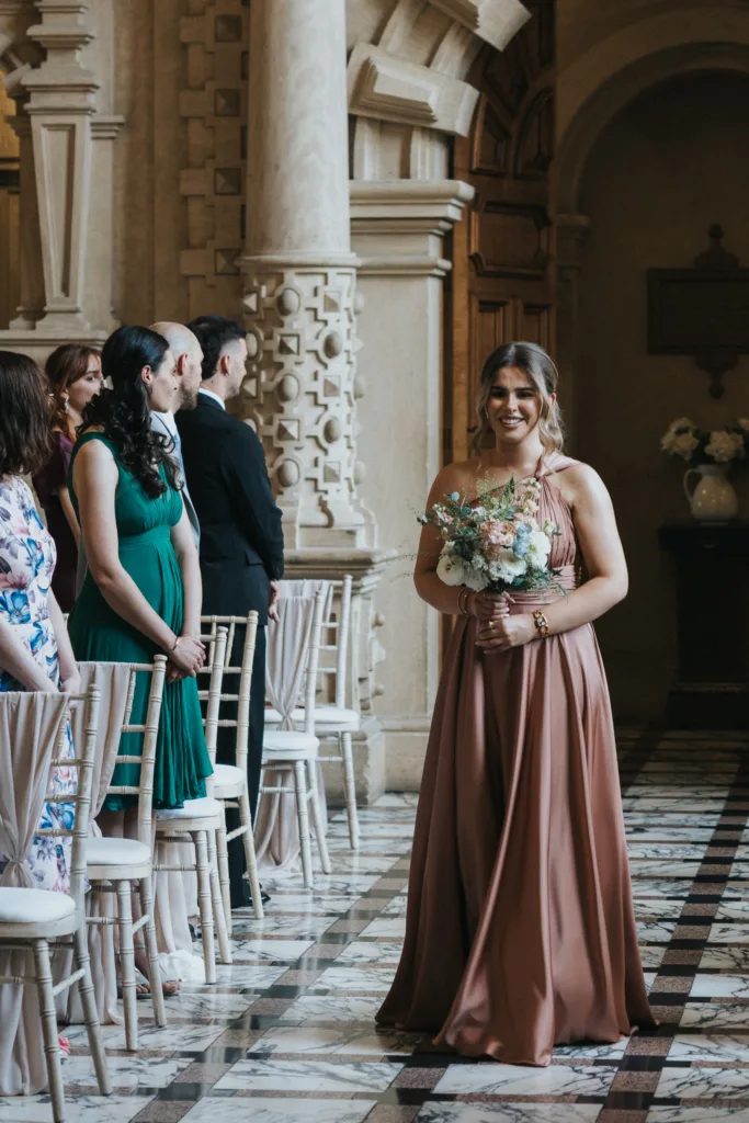 A smiling woman in a blush pink dress holds a bouquet whilst walking down an aisle lined with white chairs at a Harlaxton Manor Wedding. Guests, some standing and some seated, watch her in an ornate hall with marble floors and stone columns. © Aimee Lince Photography, Lincolnshire photographer