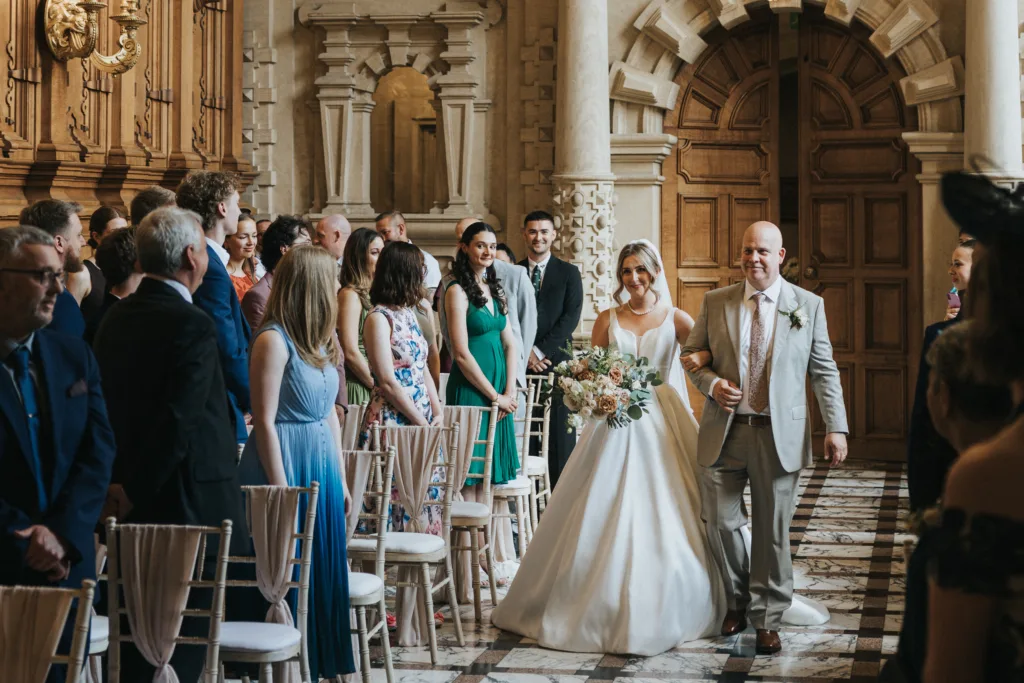 A bride in a white dress, holding a bouquet, walks down the aisle on her father’s arm at a Harlaxton Manor Wedding. Guests stand in formal attire as elegant wooden doors and classical architecture create a breathtaking backdrop. © Aimee Lince Photography, Lincolnshire photographer