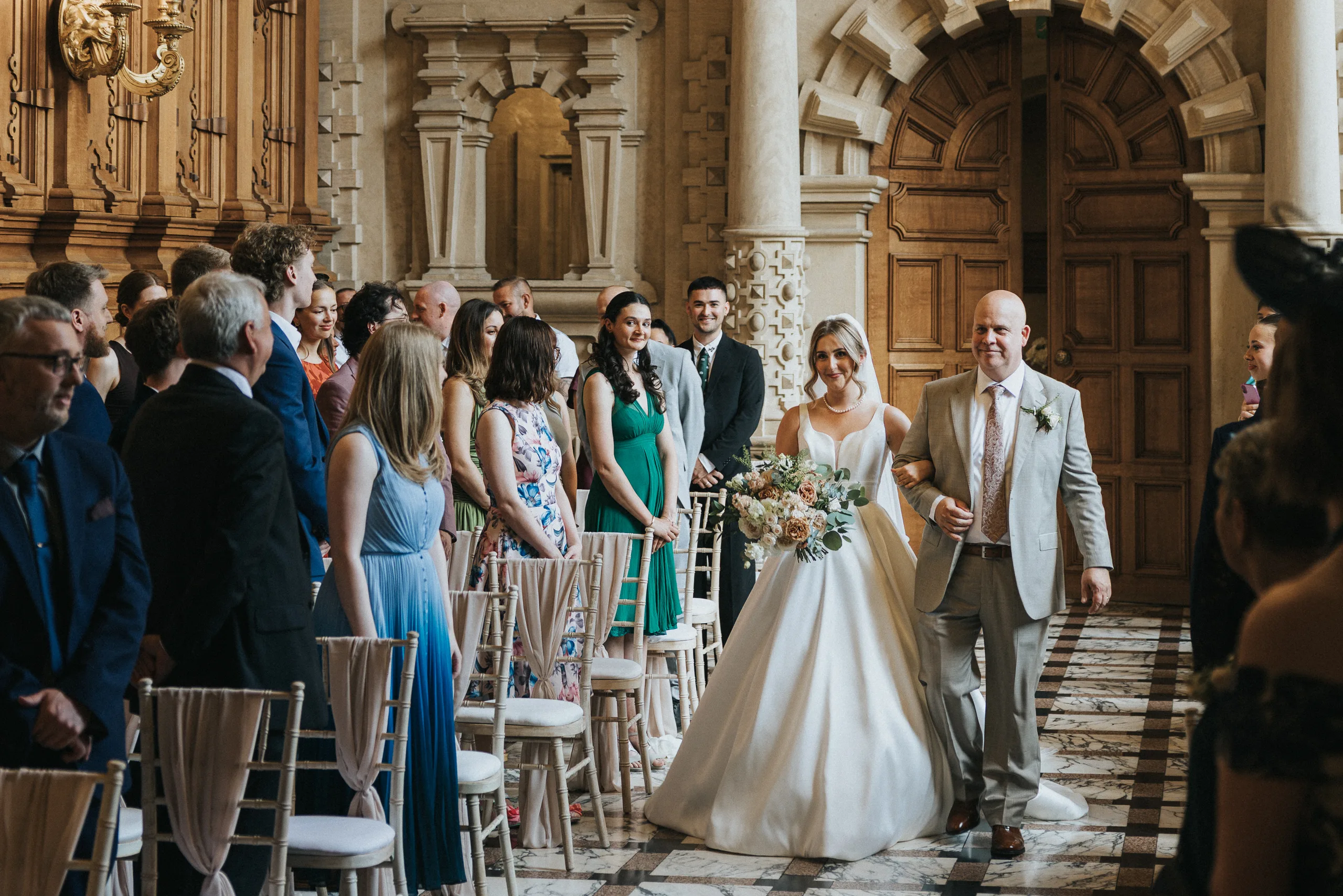 A bride in a white dress, holding a bouquet, walks down the aisle on her father’s arm at a Harlaxton Manor Wedding. Guests stand in formal attire as elegant wooden doors and classical architecture create a breathtaking backdrop. © Aimee Lince Photography, Lincolnshire photographer
