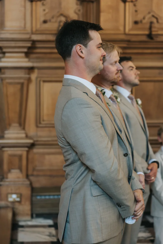 Three men in light grey suits and pale ties stand side by side in a wood-panelled room at Harlaxton Manor Wedding, hands clasped as they await the formal event. Ornate wooden panelling fills the background, setting an elegant tone. © Aimee Lince Photography, Lincolnshire photographer