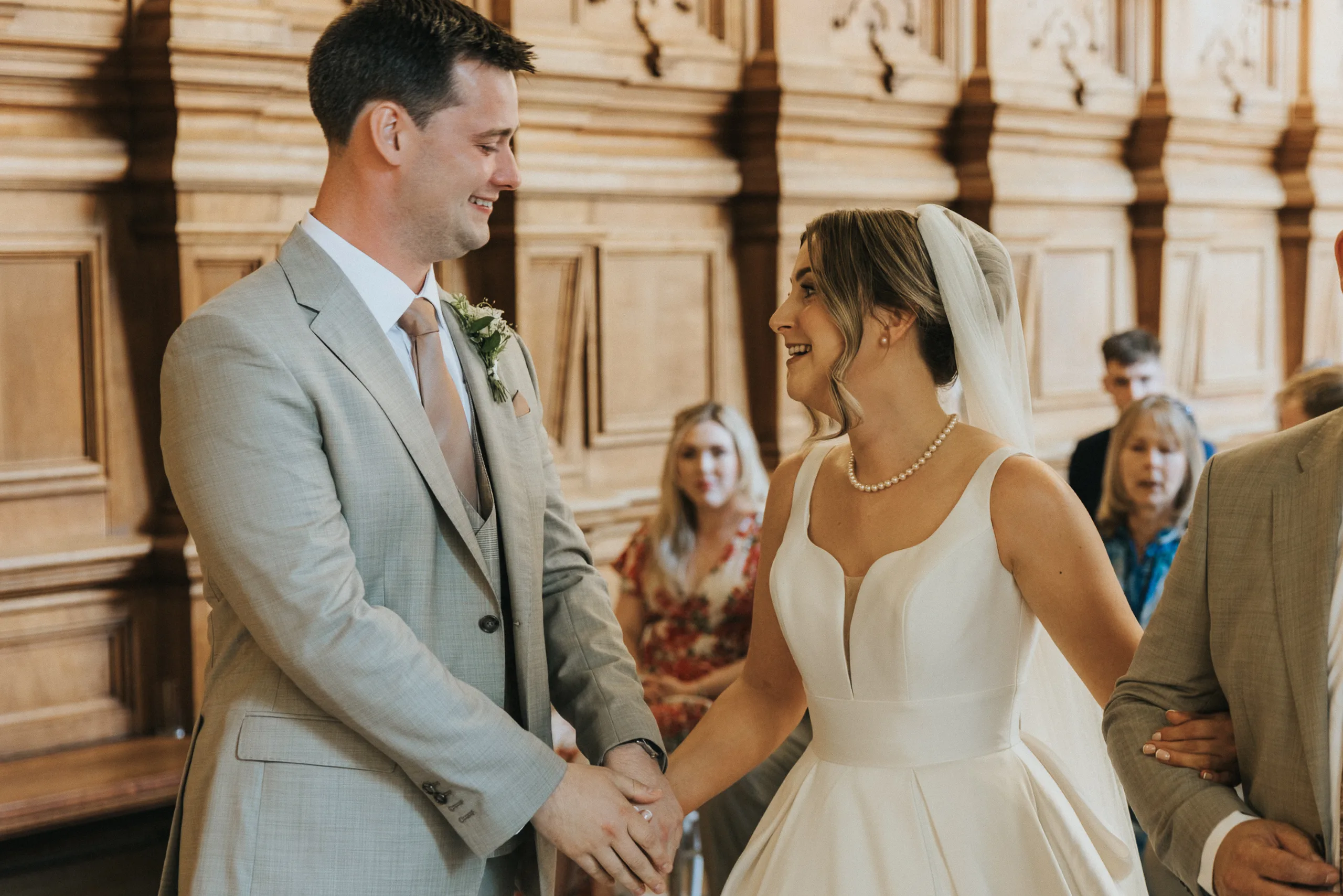 A bride in a white gown and veil holds hands and smiles at a groom in a light grey suit with a buttonhole. They stand in a wood-panelled room at their Harlaxton Manor Wedding, with guests seated behind them, witnessing the ceremony. © Aimee Lince Photography, Lincolnshire photographer