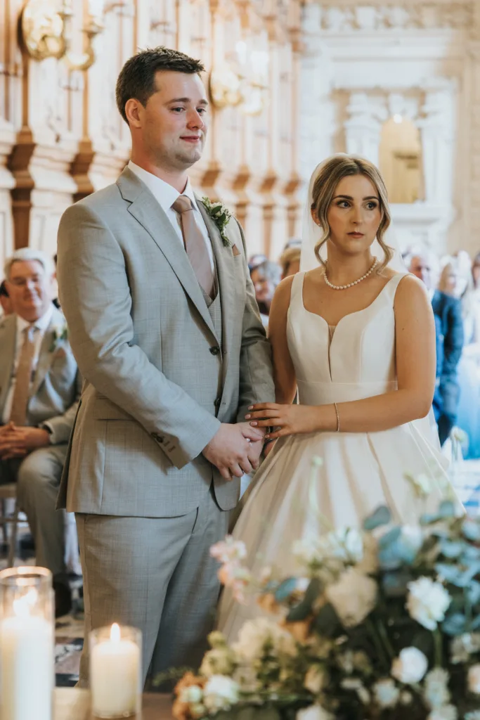 A bride in a white gown and pearl necklace stands beside a groom in a light grey suit and pink tie. They hold hands, appearing emotional, at their Harlaxton Manor wedding as guests look on in an ornate, light-filled room adorned with flowers and candles. © Aimee Lince Photography, Lincolnshire photographer