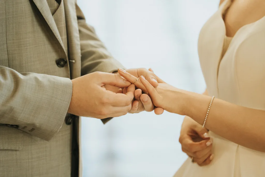 A close-up of two people in formal attire exchanging rings at a Harlaxton Manor Wedding. One, in a light grey suit, gently places a ring on the other's finger, dressed in cream with a bracelet. The softly blurred background highlights the moment's intimacy. © Aimee Lince Photography, Lincolnshire photographer