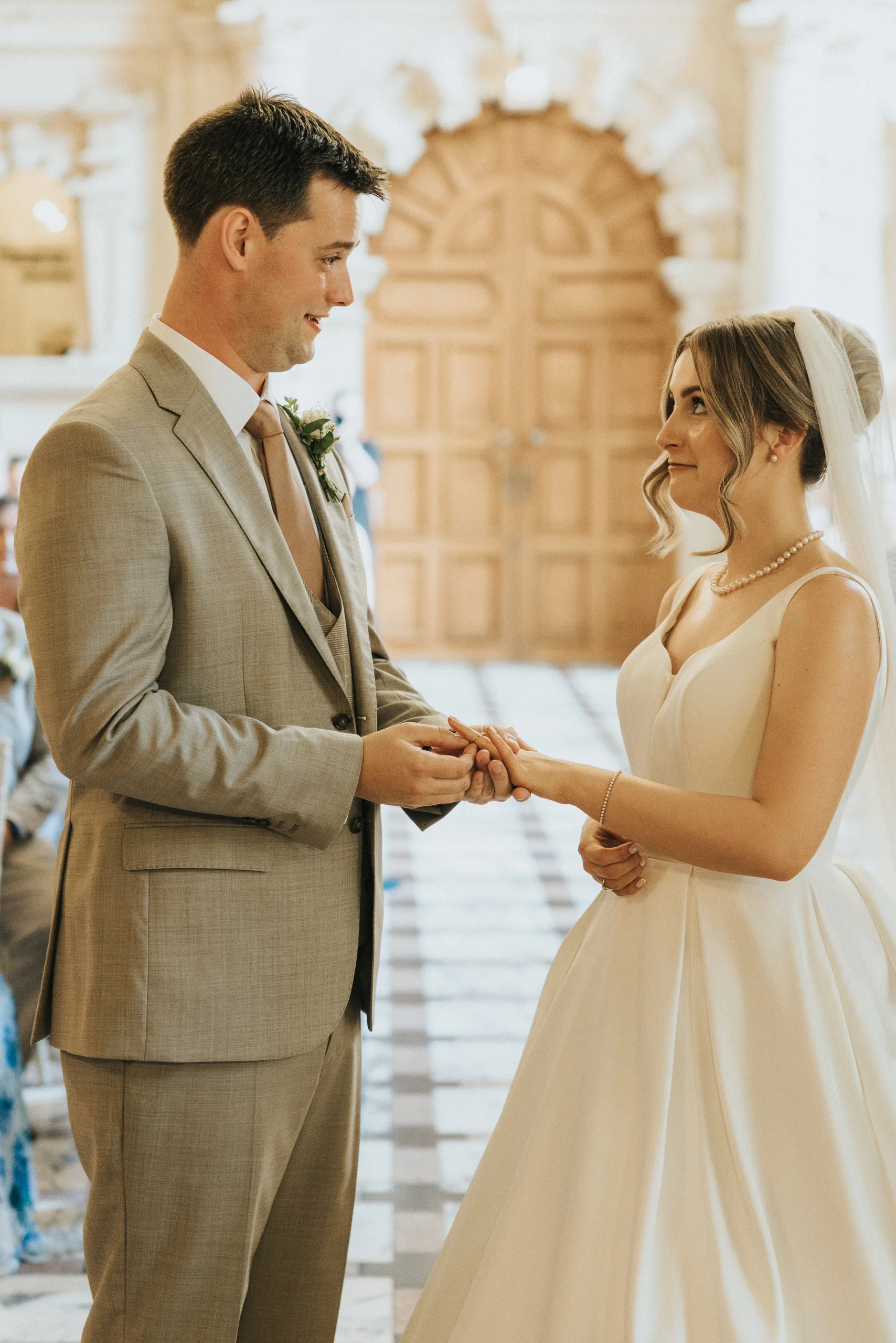 A groom in a light tan suit places a ring on the bride’s finger. The bride, in a white gown with veil and pearls, smiles as they wed inside a bright, elegant hall at their Harlaxton Manor Wedding with arched wooden doors and chequered marble floors. © Aimee Lince Photography, Lincolnshire photographer