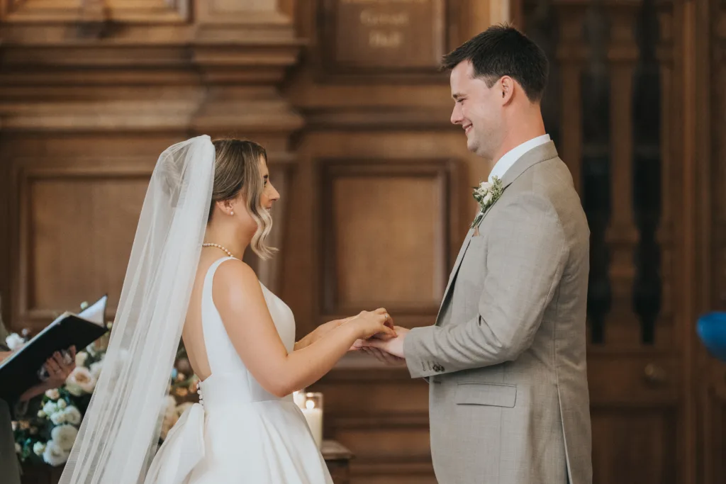 A bride in a white dress and veil exchanges rings with a groom in a light grey suit during an indoor Harlaxton Manor Wedding. They are smiling at each other, holding hands, surrounded by wood-panelled walls and floral arrangements. © Aimee Lince Photography, Lincolnshire photographer