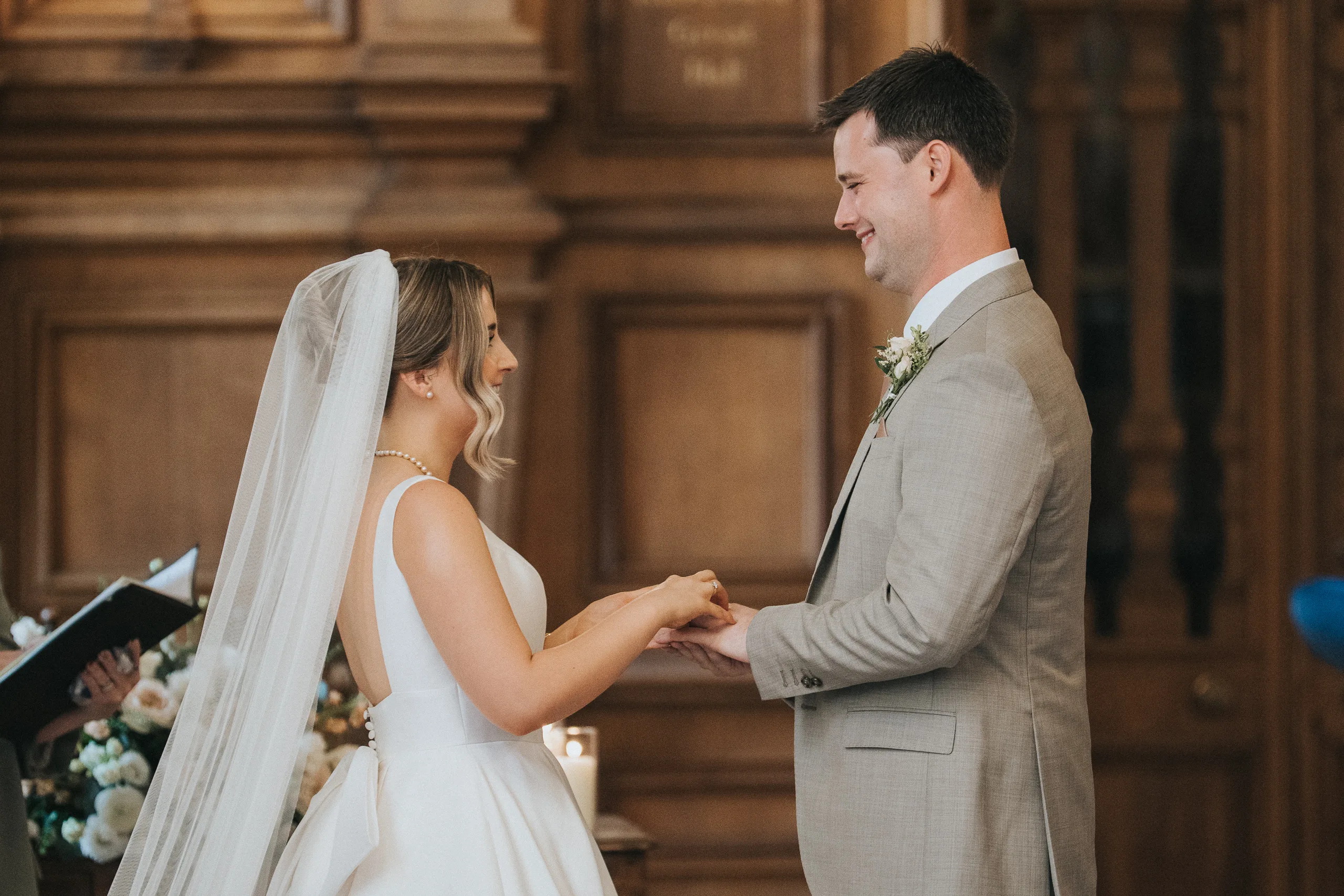 A bride in a white dress and veil exchanges rings with a groom in a light grey suit during an indoor Harlaxton Manor Wedding. They are smiling at each other, holding hands, surrounded by wood-panelled walls and floral arrangements. © Aimee Lince Photography, Lincolnshire photographer