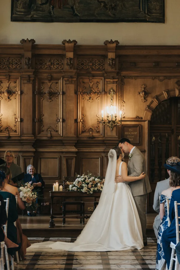 A bride and groom share a kiss at their indoor Harlaxton Manor wedding, standing before a wooden panelled wall. Guests sit on either side, some smiling, while candles and floral arrangements create a warm and elegant atmosphere. © Aimee Lince Photography, Lincolnshire photographer