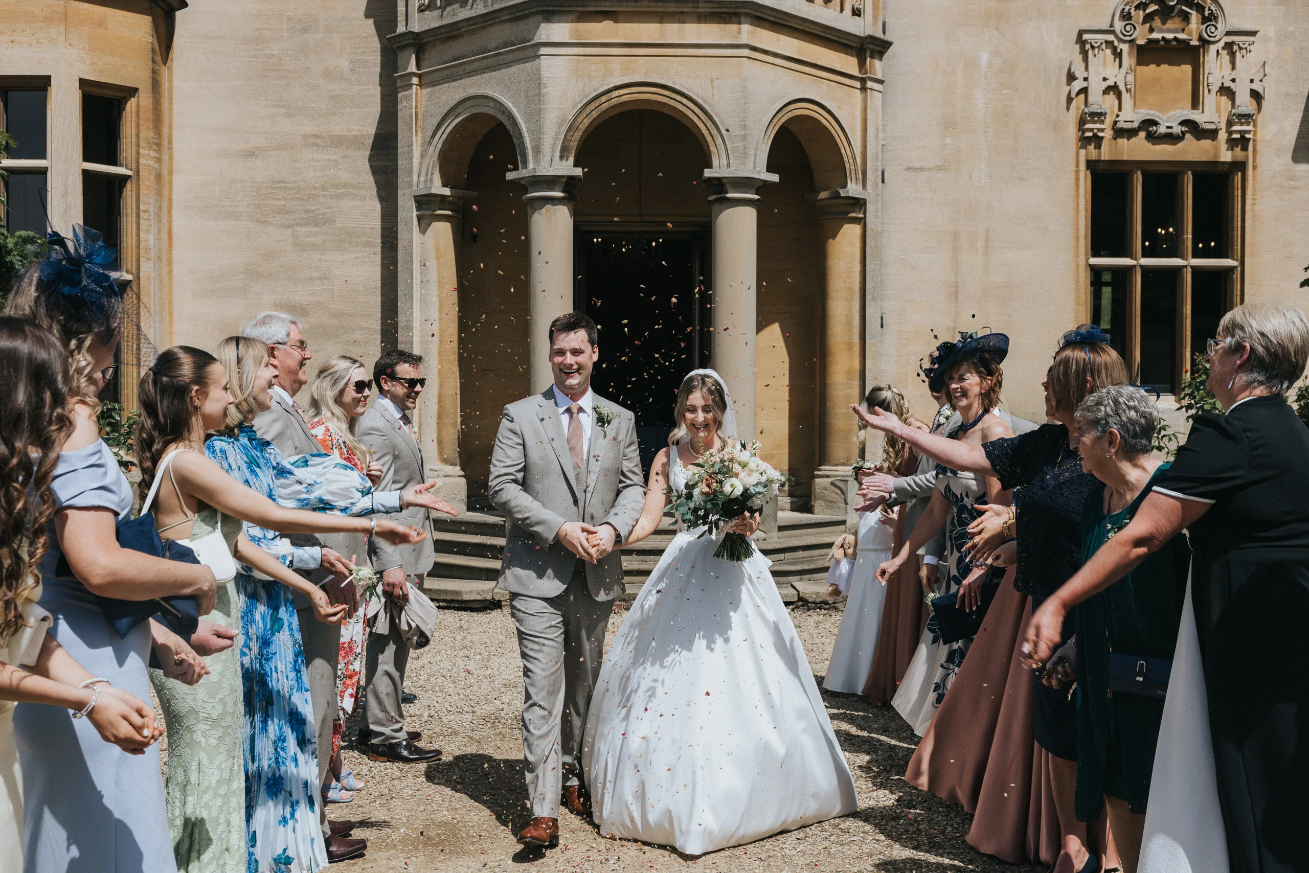 A bride and groom walk hand in hand, smiling, between two rows of guests outside the grand stone façade of a Harlaxton Manor wedding. The bride holds a bouquet; confetti fills the air as guests celebrate under the sun. © Aimee Lince Photography, Lincolnshire photographer