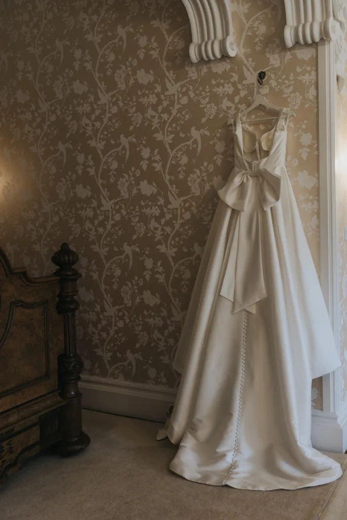 A white satin wedding dress with a large bow on the back and a row of covered buttons hangs on an ornate hook against beige, floral-patterned wallpaper—perfect for an elegant Harlaxton Manor Wedding. Part of a carved wooden bedframe appears on the left. © Aimee Lince Photography, Lincolnshire photographer