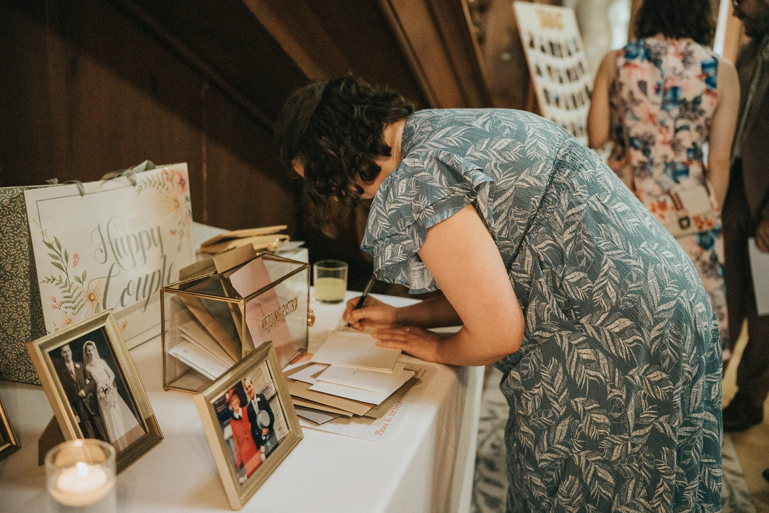At a Harlaxton Manor Wedding, a woman in a blue patterned dress bends over a table, writing on a card. The table holds framed photos, a candle, drink, glass card box, and "Happy Couple" sign. Guests and photo display fill the background. © Aimee Lince Photography, Lincolnshire photographer