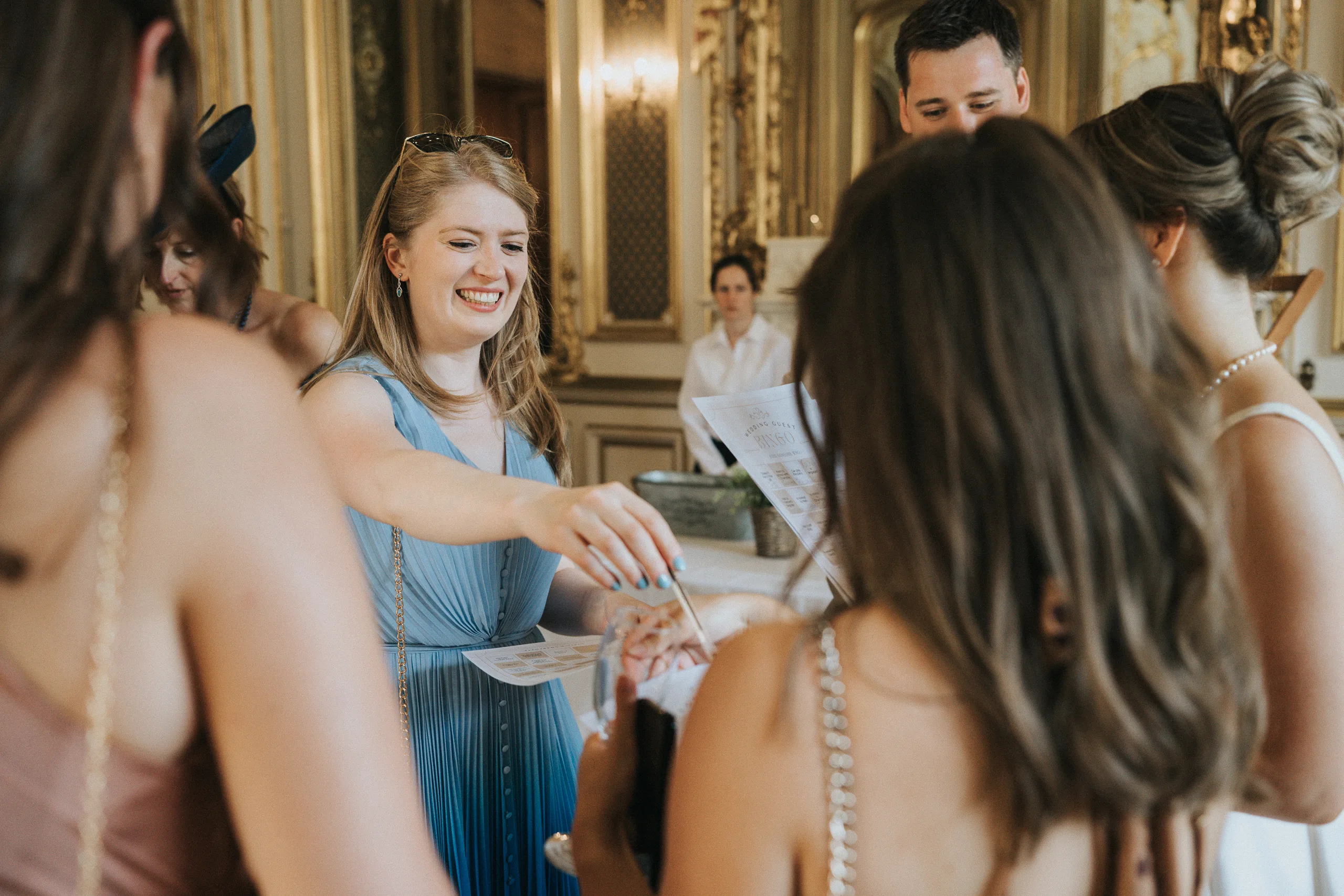 A smiling woman in a light blue dress interacts with several people indoors at an elegant Harlaxton Manor wedding. She reaches towards someone while holding paper in the ornate room, which features gold accents, mirrors, and a seated figure in the background. © Aimee Lince Photography, Lincolnshire photographer