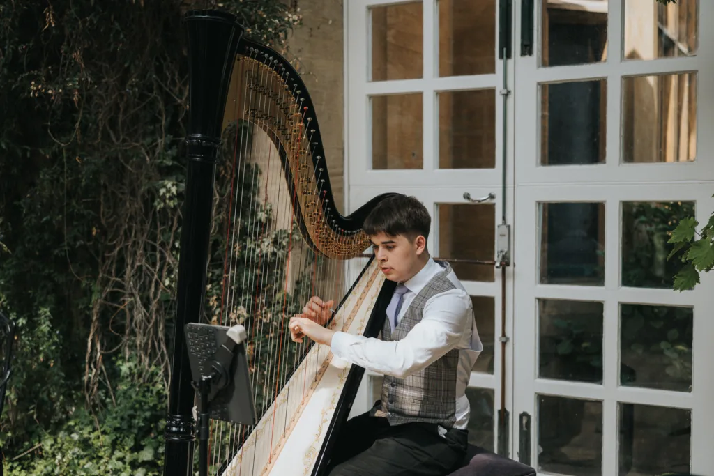 A young man in a waistcoat, shirt, and tie plays a large black harp outdoors at a Harlaxton Manor wedding. He is seated, focused on the strings as soft natural light and leafy greenery frame the elegant glass doors behind him. © Aimee Lince Photography, Lincolnshire photographer