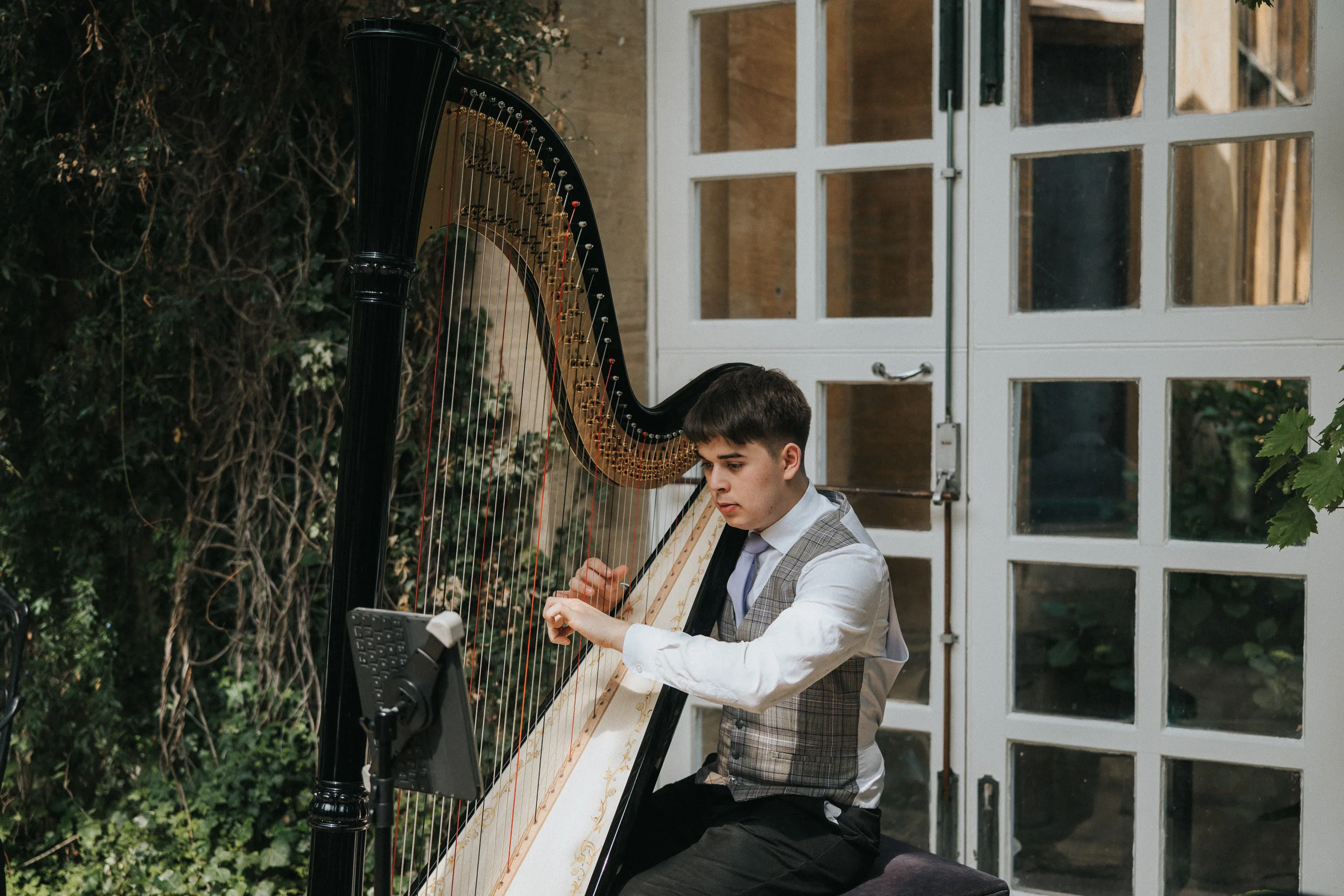 A young man in a waistcoat, shirt, and tie plays a large black harp outdoors at a Harlaxton Manor wedding. He is seated, focused on the strings as soft natural light and leafy greenery frame the elegant glass doors behind him. © Aimee Lince Photography, Lincolnshire photographer