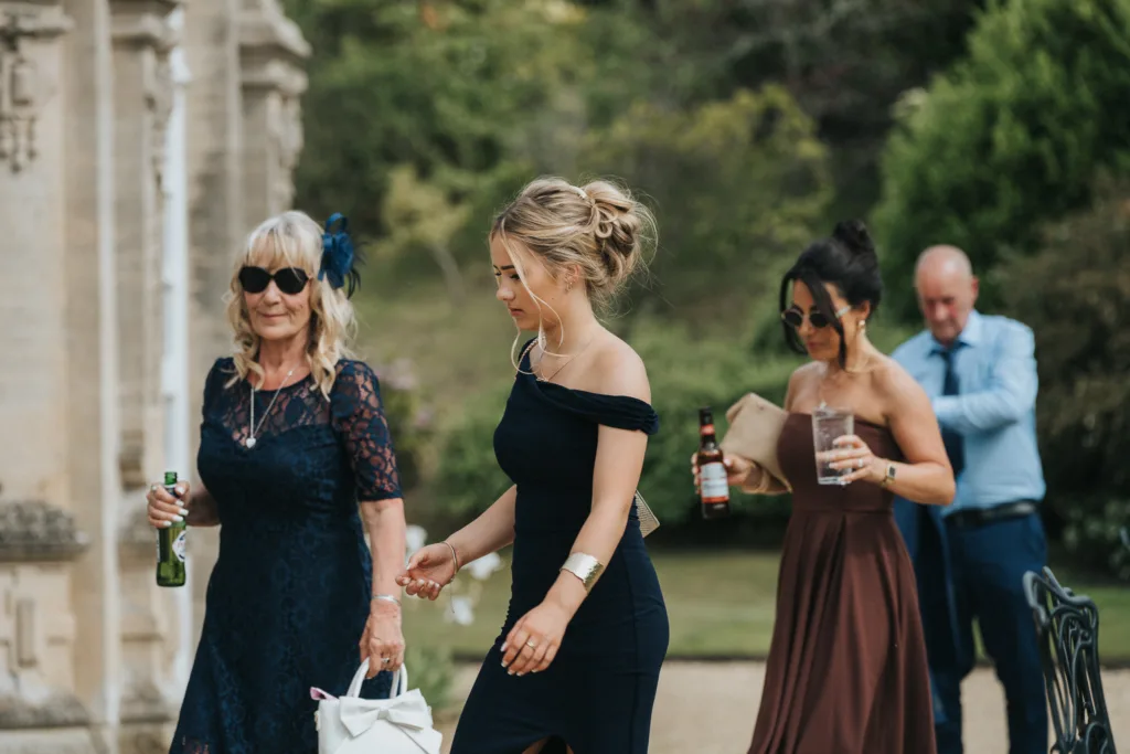 Four people dressed formally walk outdoors by a stone building at a Harlaxton Manor wedding. Two women in front wear dark dresses; one older with sunglasses and a handbag, the other younger with blonde hair up. Behind, a woman and man hold drinks amid lush greenery. © Aimee Lince Photography, Lincolnshire photographer