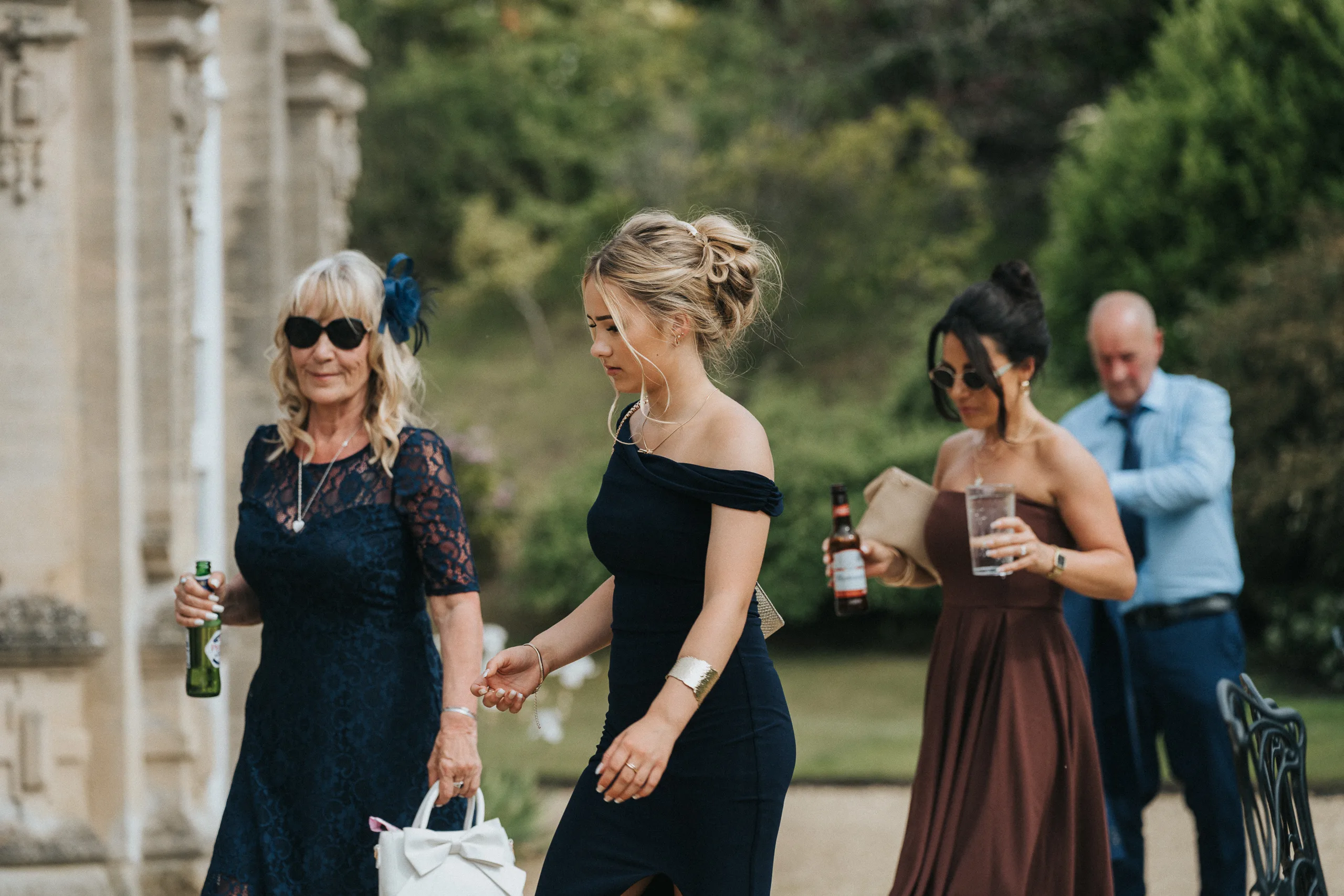 Four people dressed formally walk outdoors by a stone building at a Harlaxton Manor wedding. Two women in front wear dark dresses; one older with sunglasses and a handbag, the other younger with blonde hair up. Behind, a woman and man hold drinks amid lush greenery. © Aimee Lince Photography, Lincolnshire photographer