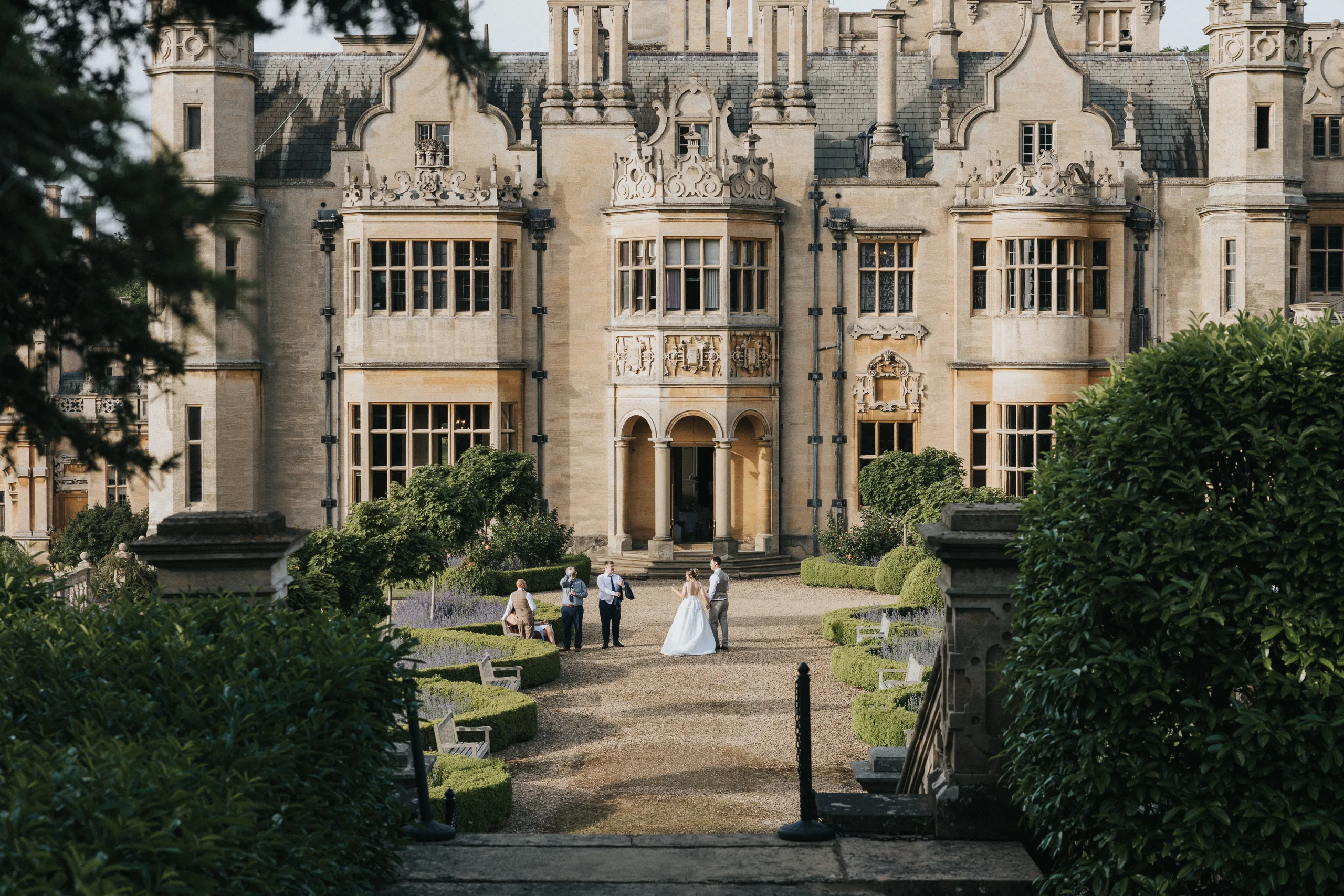 A bride and groom stand together in a landscaped garden, flanked by three guests, in front of the grand, historic stone façade of Harlaxton Manor. Lush greenery and trimmed hedges beautifully frame this elegant Harlaxton Manor wedding scene. © Aimee Lince Photography, Lincolnshire photographer