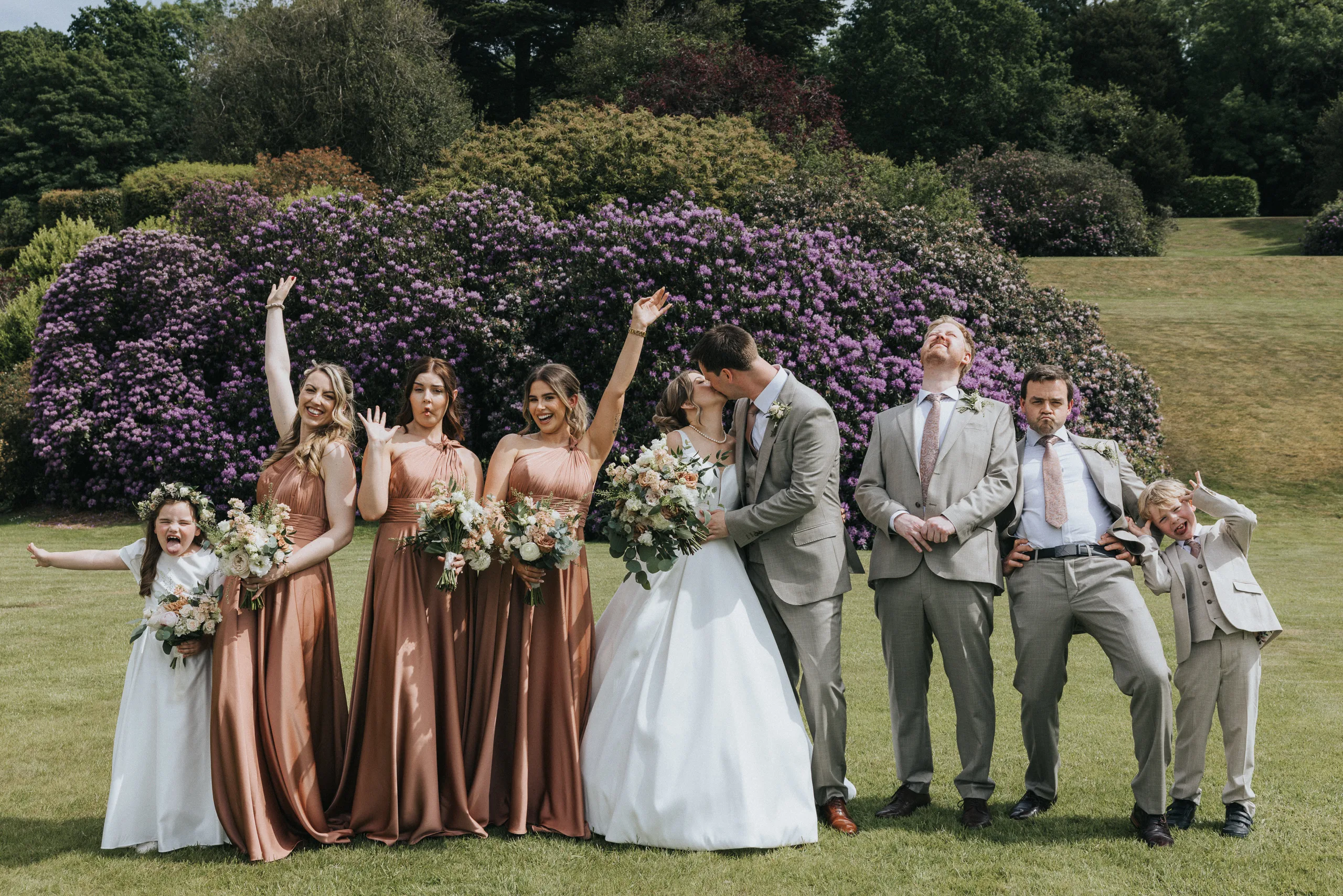 A wedding party poses on grass before purple flowering bushes at a Harlaxton Manor Wedding. The bride and groom kiss centre, bridesmaids in copper dresses and groomsmen in beige suits cheer whilst children at each end strike joyful poses. © Aimee Lince Photography, Lincolnshire photographer