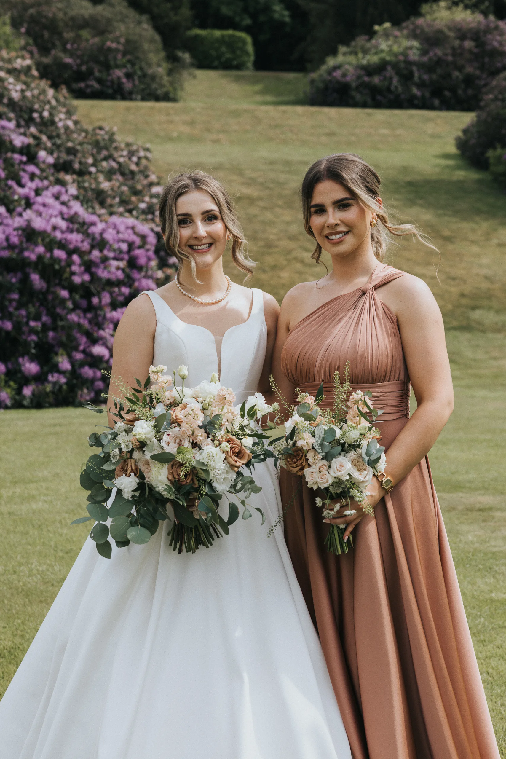 Two women stand side by side outside on a lawn at a Harlaxton Manor Wedding. One wears a white wedding dress and pearl necklace, the other a mauve bridesmaid dress. Both hold bouquets, with blooming purple shrubs and trees in the background. © Aimee Lince Photography, Lincolnshire photographer