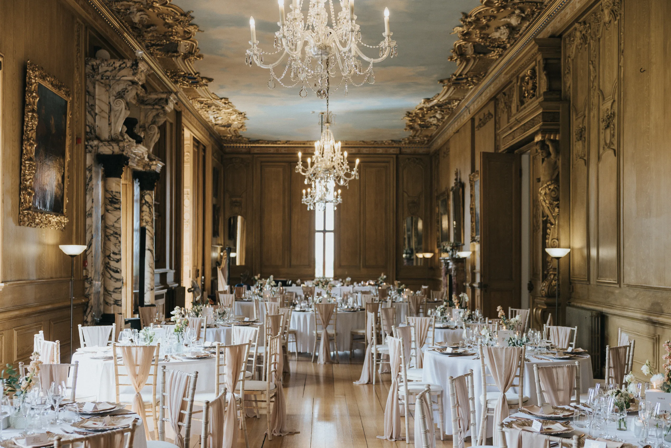 Elegant banqueting hall with ornate gold walls, chandeliers, and a painted ceiling—perfect for a Harlaxton Manor Wedding. Tables with white linen and floral centrepieces are neatly arranged, while classic paintings and large windows add grandeur to the setting. © Aimee Lince Photography, Lincolnshire photographer
