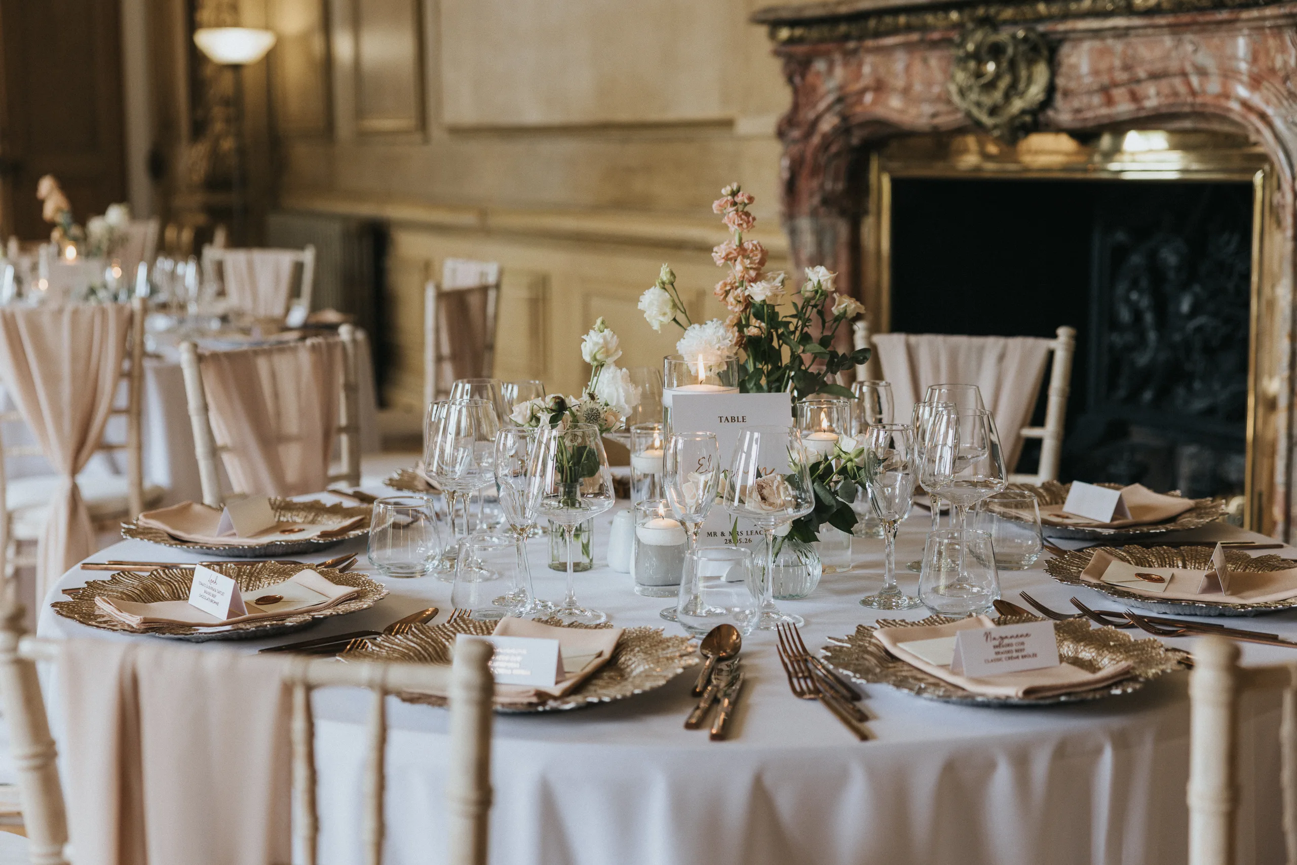 Elegant round dining tables set for a formal event, featuring white tablecloths, gold-rimmed plates, glassware, floral centrepieces, and candles. In the background, a carved fireplace evokes Harlaxton Manor Wedding sophistication. © Aimee Lince Photography, Lincolnshire photographer