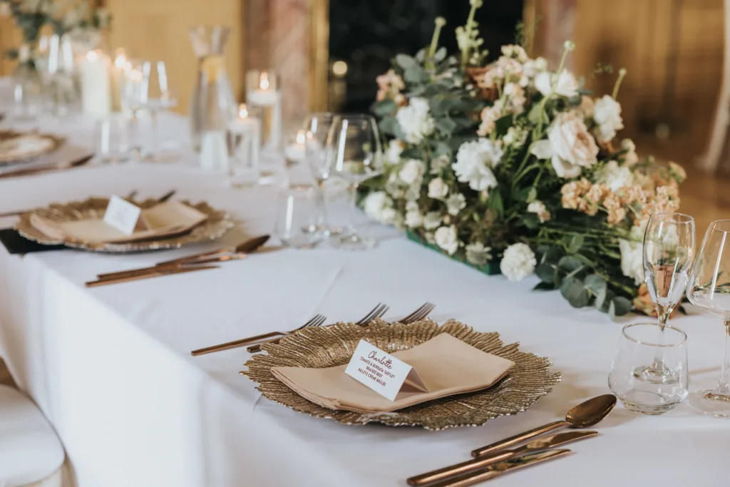 Elegant dining table set for a formal Harlaxton Manor Wedding with gold charger plates, beige serviettes, name cards, glassware, and a floral centrepiece of white and peach blooms on a white tablecloth. Candles and jugs add charm in the background. © Aimee Lince Photography, Lincolnshire photographer