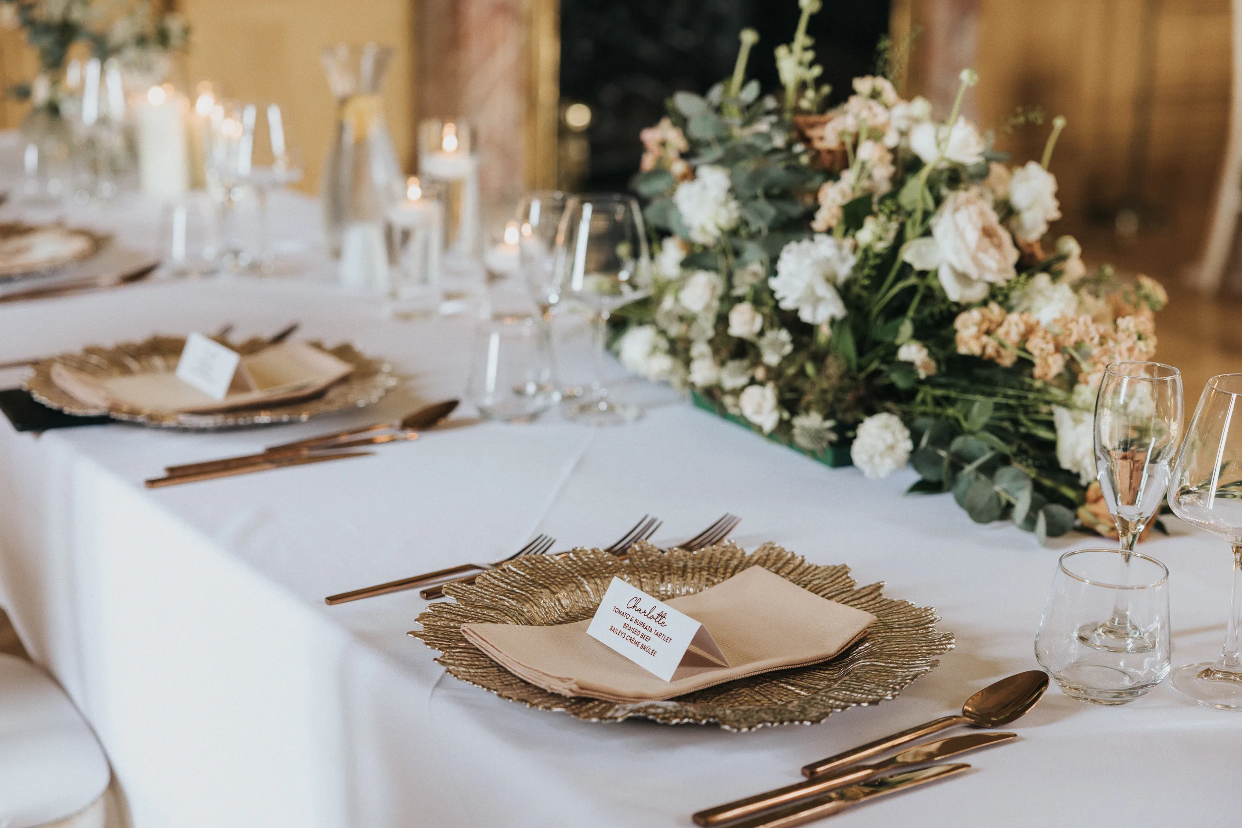Elegant dining table set for a formal Harlaxton Manor Wedding with gold charger plates, beige serviettes, name cards, glassware, and a floral centrepiece of white and peach blooms on a white tablecloth. Candles and jugs add charm in the background. © Aimee Lince Photography, Lincolnshire photographer