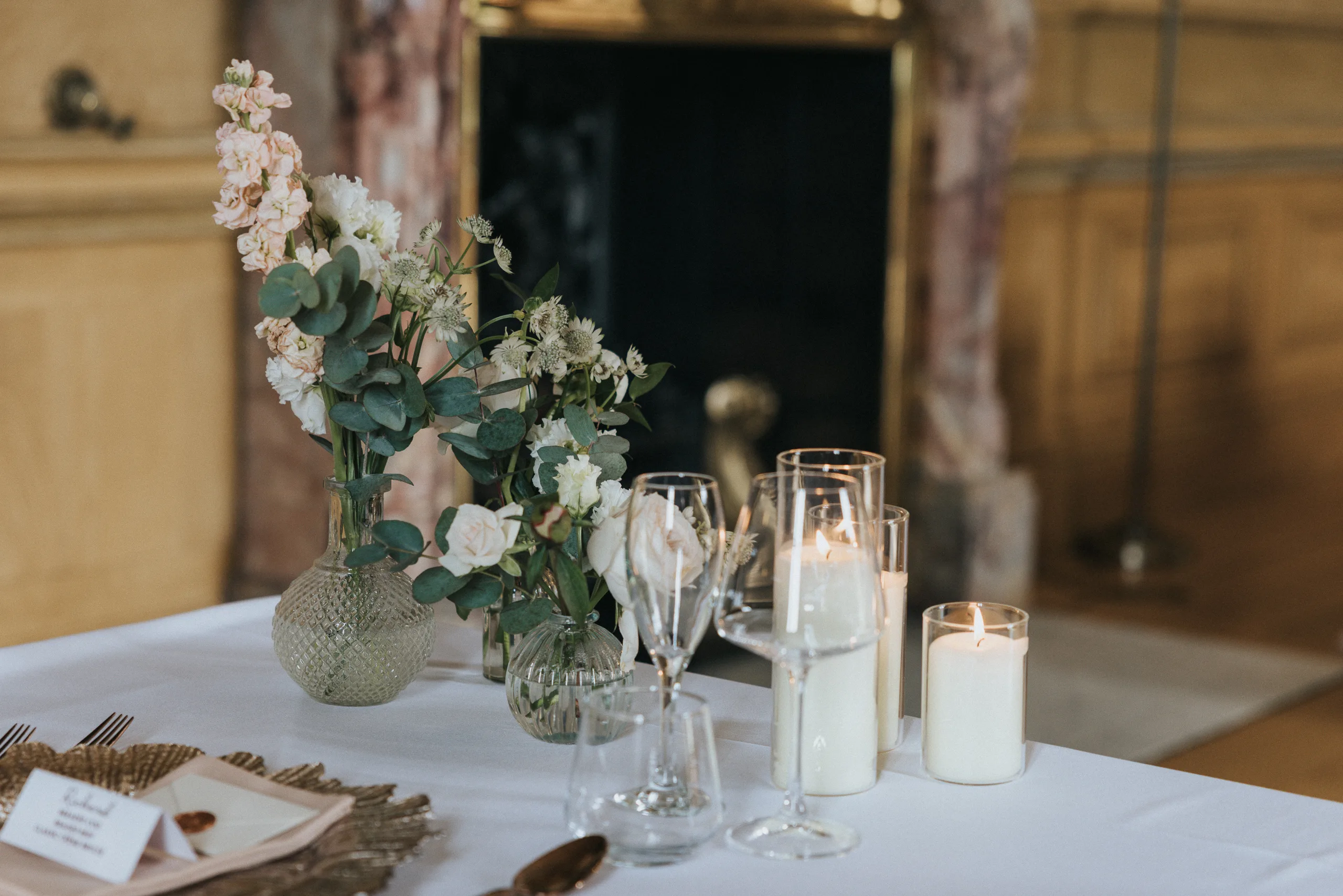 A white tablecloth holds glass vases with white flowers and greenery, three lit pillar candles, empty wine glasses, and plates with serviettes—perfect for an elegant Harlaxton Manor wedding in a warmly lit room with a marble and brass-detailed fireplace. © Aimee Lince Photography, Lincolnshire photographer
