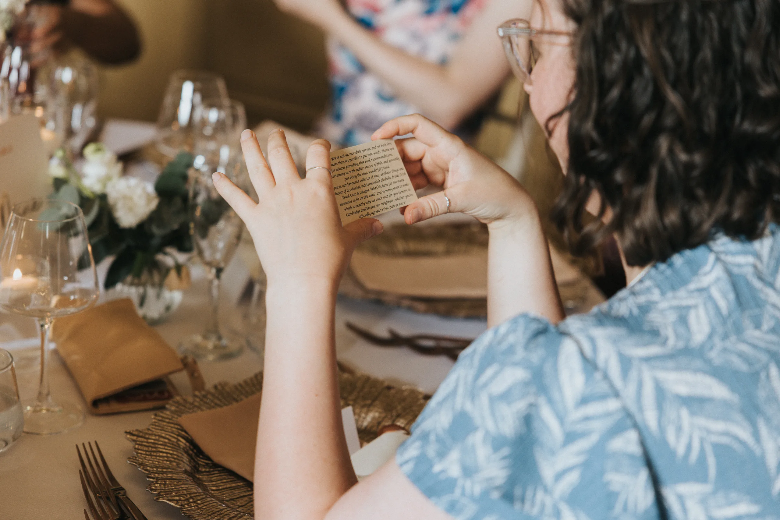 A person with wavy brown hair and glasses, wearing a blue patterned shirt, holds a small card with text at a decorated Harlaxton Manor Wedding table set for a meal, complete with wine glasses, plates, cutlery, and a floral centrepiece. © Aimee Lince Photography, Lincolnshire photographer