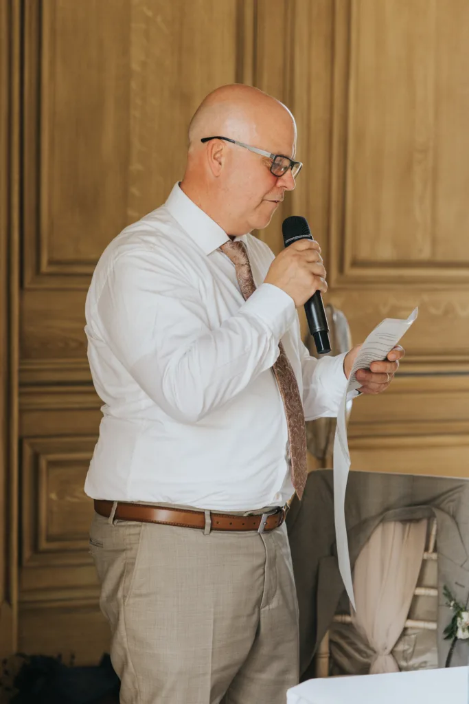 A bald man with glasses, wearing a white shirt, patterned tie, and beige trousers, stands indoors at a Harlaxton Manor wedding holding a microphone and a sheet of paper, as he reads or speaks. Wooden panelled walls are in the background. © Aimee Lince Photography, Lincolnshire photographer