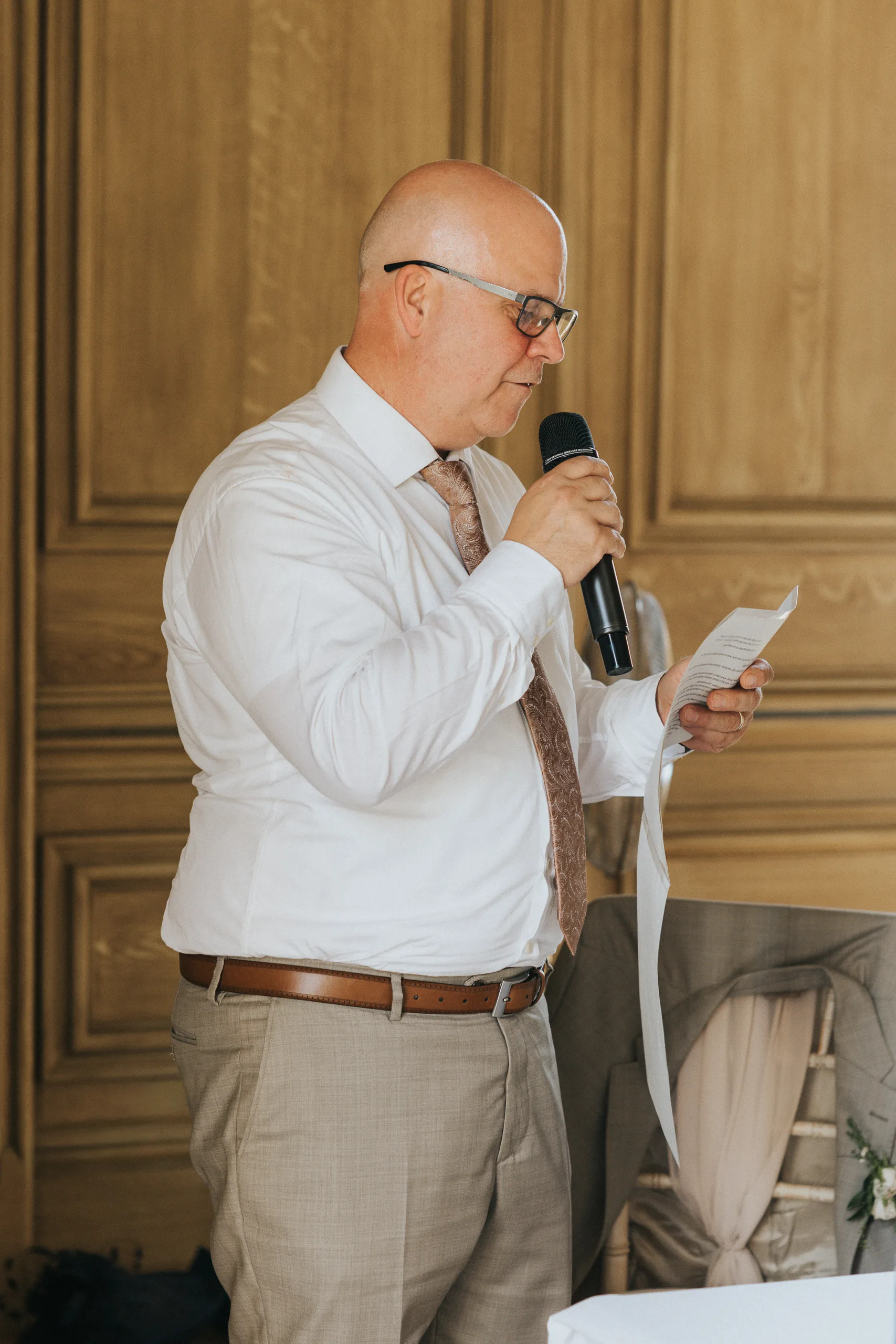 A bald man with glasses, wearing a white shirt, patterned tie, and beige trousers, stands indoors at a Harlaxton Manor wedding holding a microphone and a sheet of paper, as he reads or speaks. Wooden panelled walls are in the background. © Aimee Lince Photography, Lincolnshire photographer