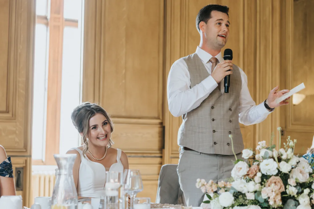 A man in a tan waistcoat and white shirt stands, holding a microphone and speech, smiling as he speaks at a Harlaxton Manor Wedding. Next to him, a woman in a white dress and pearl necklace sits, smiling up at him at their flower-adorned table in a wood-panelled room. © Aimee Lince Photography, Lincolnshire photographer