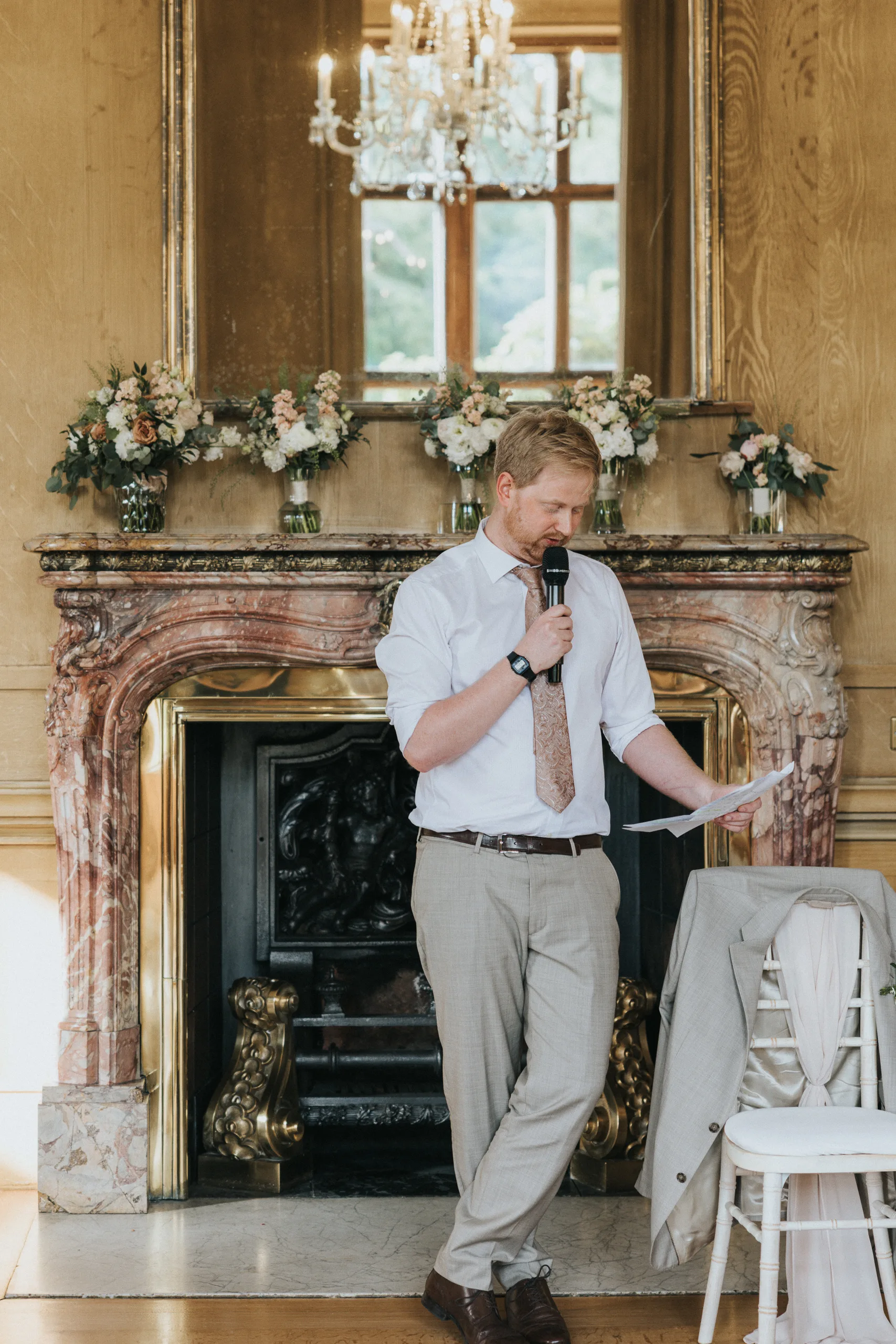 A man with fair hair and a beard stands in front of a grand marble fireplace at a Harlaxton Manor Wedding, holding a microphone and reading from a piece of paper. He wears a white shirt, patterned tie, and beige trousers amid elegant flowers and a chandelier. © Aimee Lince Photography, Lincolnshire photographer