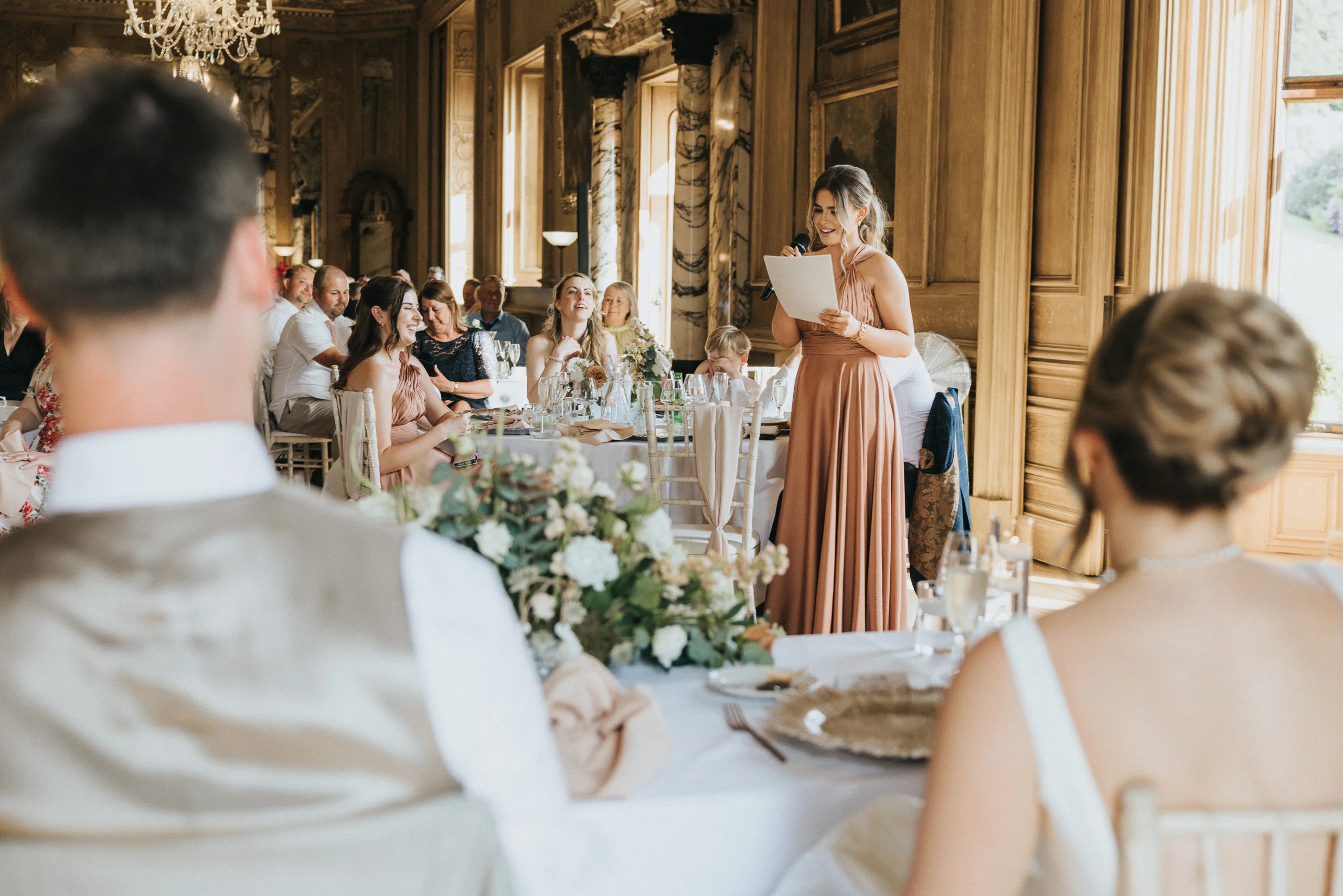 A woman in a peach dress stands, reading from a paper, giving a speech at a Harlaxton Manor wedding in an elegant, ornate room. Guests seated at tables with white tablecloths and floral centrepieces listen attentively. © Aimee Lince Photography, Lincolnshire photographer