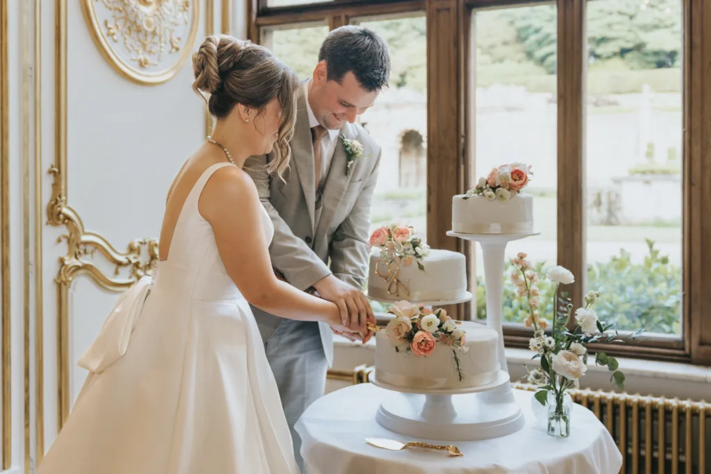 A bride in a white gown and groom in a light grey suit smile as they cut a white, two-tier wedding cake adorned with pink and peach flowers at their elegant Harlaxton Manor wedding, surrounded by gold accents and large windows. © Aimee Lince Photography, Lincolnshire photographer