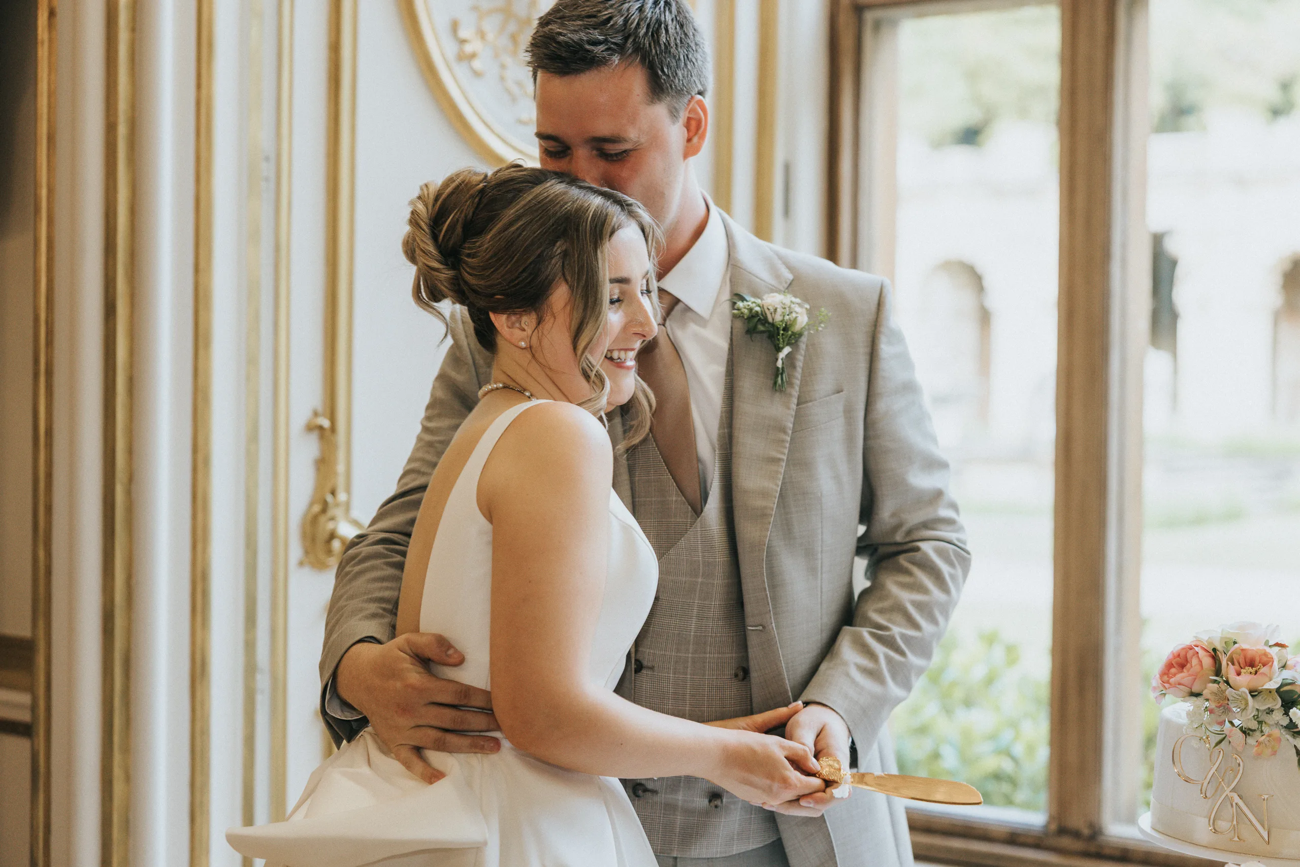 A bride in a white dress and a groom in a light grey suit smile as they cut their wedding cake at their Harlaxton Manor Wedding. The groom embraces the bride from behind and kisses her head, surrounded by ornate gold details and soft natural light. © Aimee Lince Photography, Lincolnshire photographer