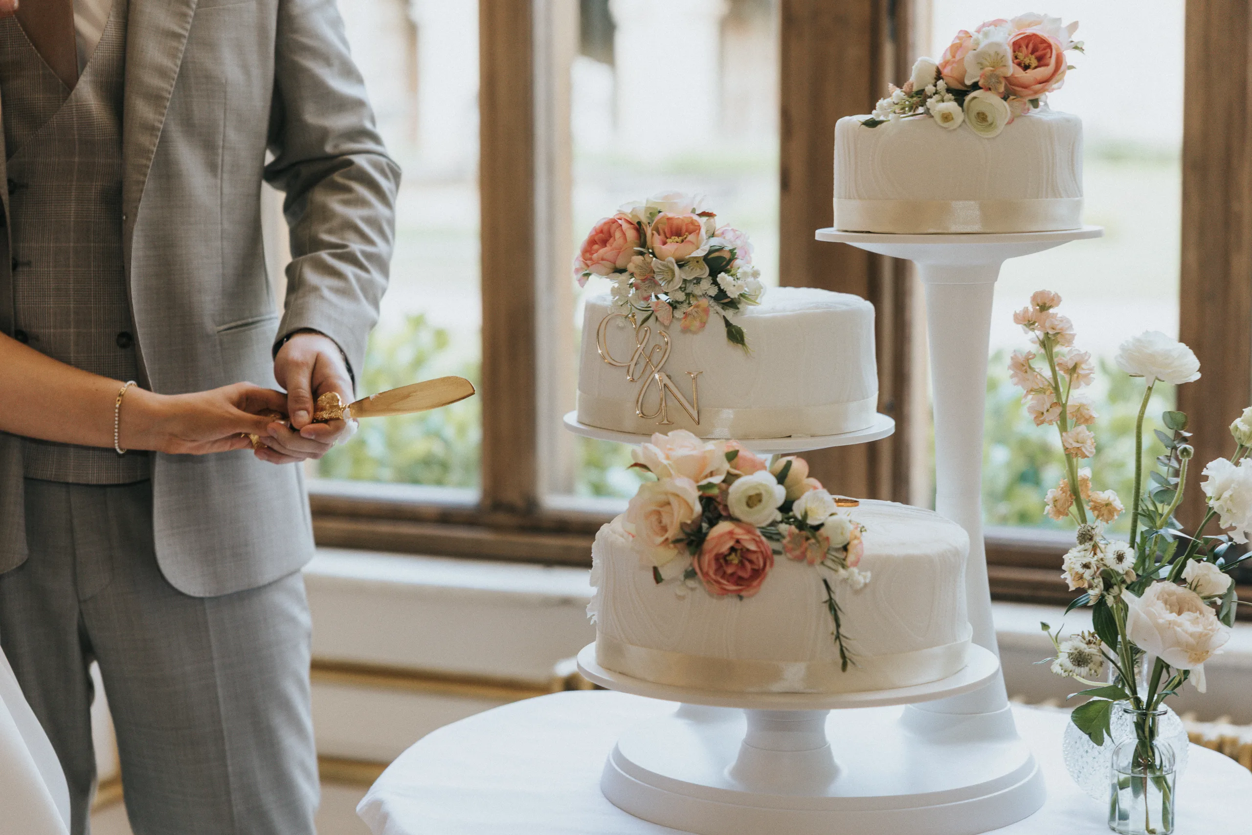A couple in formal attire are about to cut a three-tiered wedding cake decorated with pink and white flowers at their Harlaxton Manor wedding. The cake sits by a bright window with floral arrangements beside it; only their hands are visible. © Aimee Lince Photography, Lincolnshire photographer