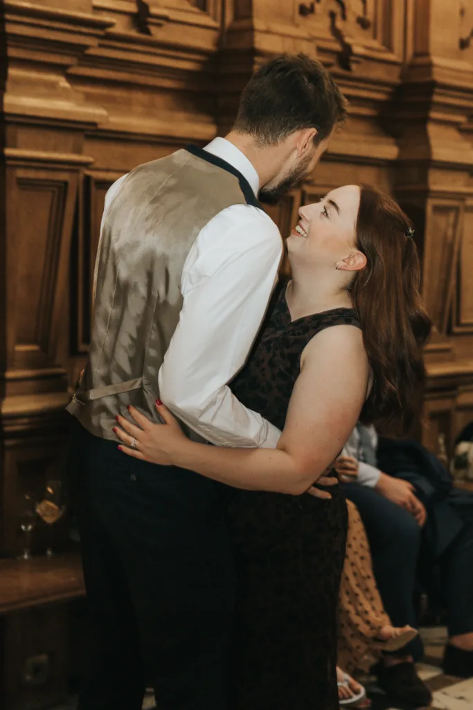 A couple embrace and smile at each other in a richly panelled wood interior at their Harlaxton Manor wedding. The man has short hair and wears a waistcoat over a white shirt; the woman, with long brown hair, is radiant in her sleeveless, patterned dress. © Aimee Lince Photography, Lincolnshire photographer