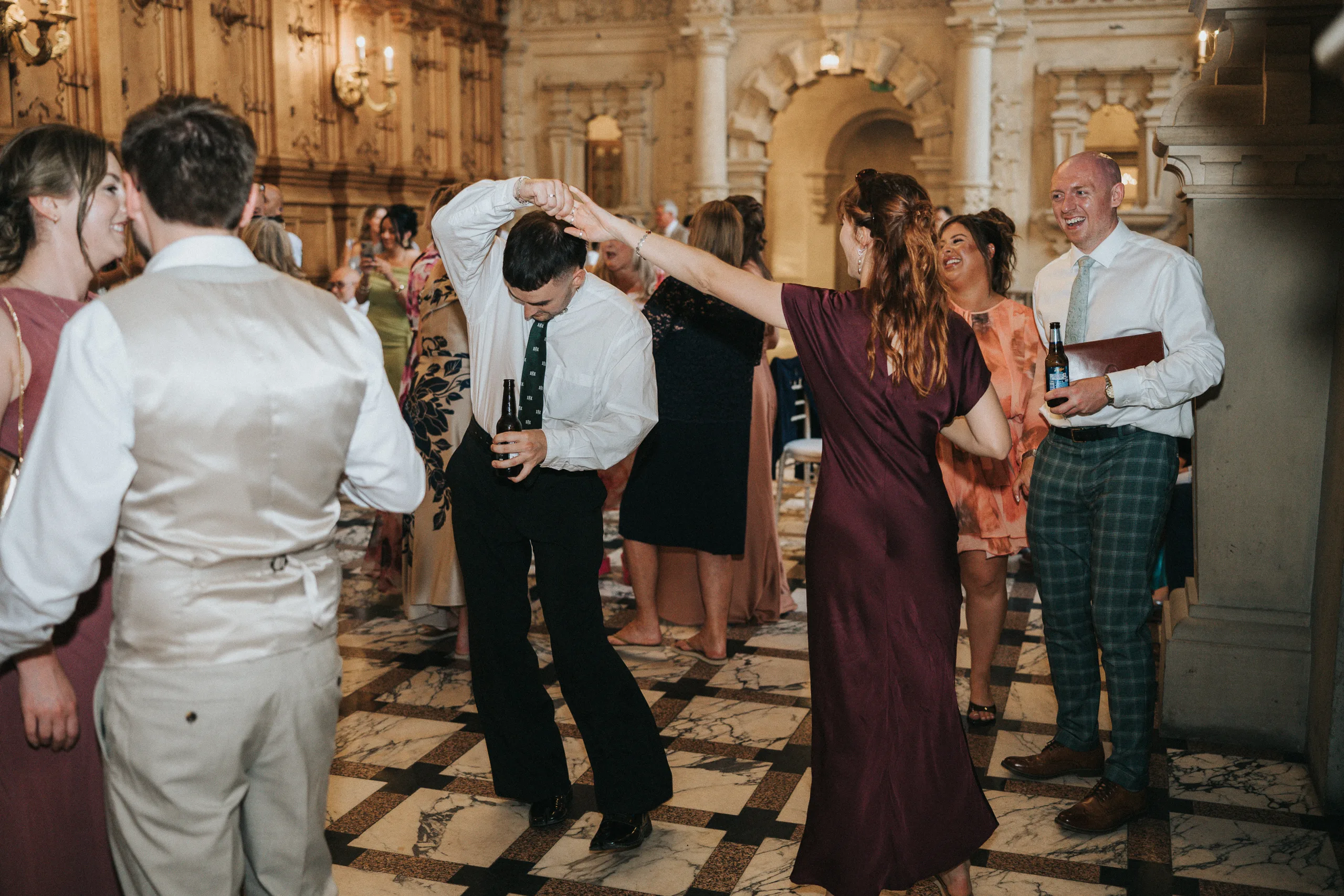 A lively group dances in an ornate hall with marble floors at a Harlaxton Manor Wedding. A woman in a maroon dress twirls a man holding a drink as other guests in formal attire smile, surrounded by arched doorways and elaborate wall carvings. © Aimee Lince Photography, Lincolnshire photographer