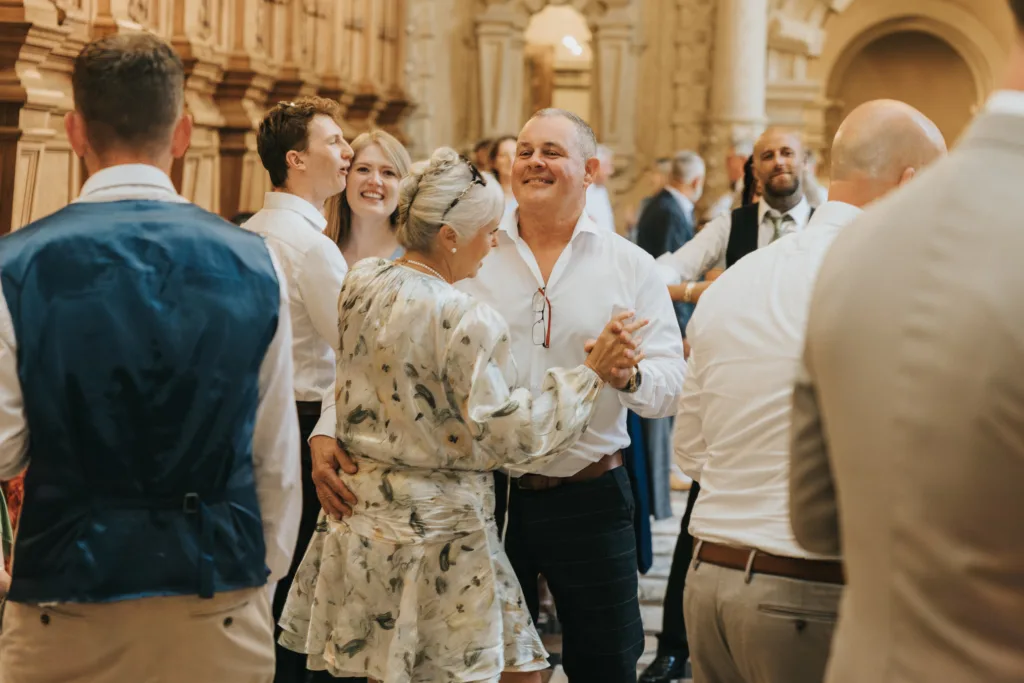 A smiling older couple dance together at a Harlaxton Manor Wedding, surrounded by guests in formal attire. The woman wears a floral dress and glasses whilst the man is in a white shirt. The elegant hall buzzes with lively conversation and joy. © Aimee Lince Photography, Lincolnshire photographer
