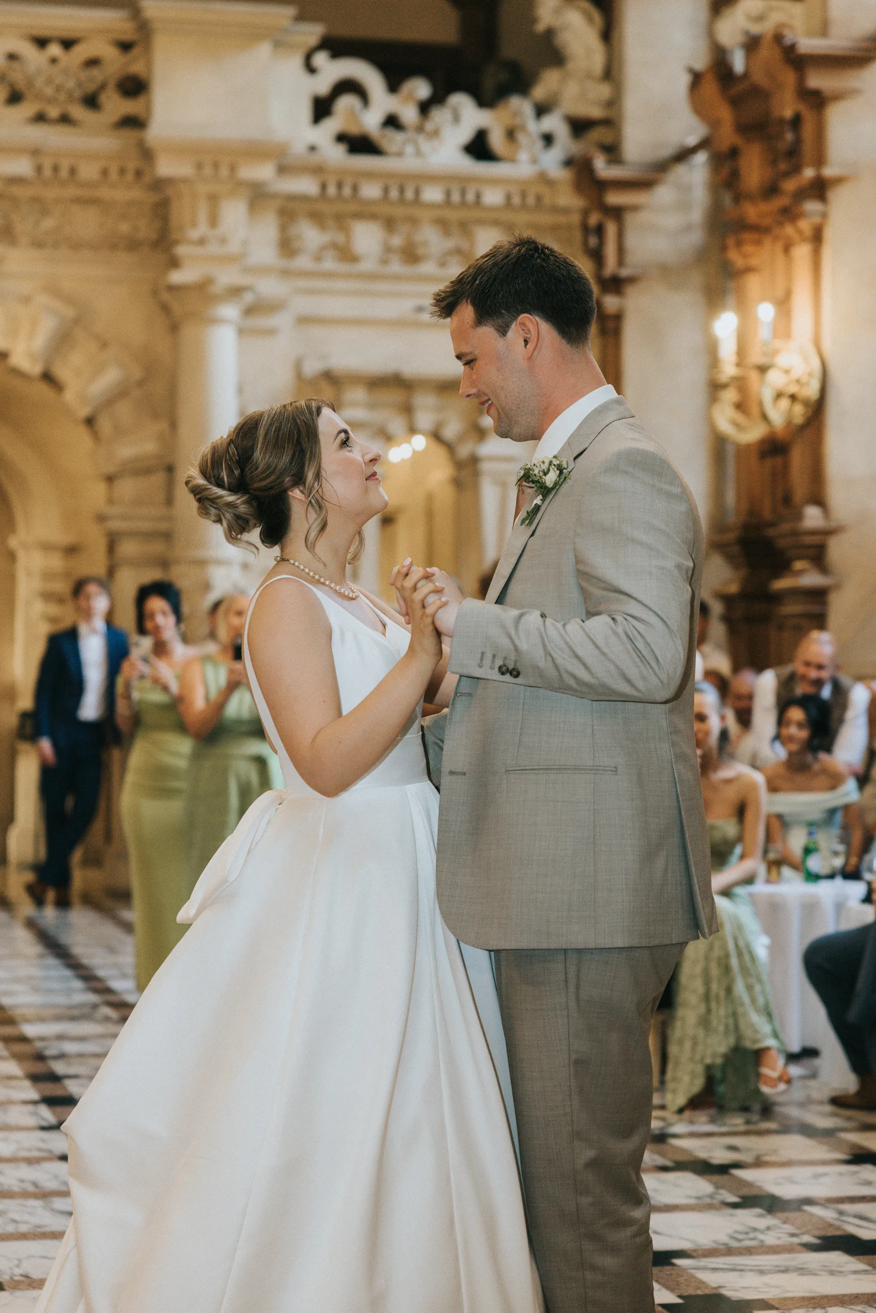 A bride in a white gown and a groom in a light grey suit share their first dance at a Harlaxton Manor Wedding. They gaze at each other, smiling, as guests watch from decorated tables amid marble floors and elegant architecture. © Aimee Lince Photography, Lincolnshire photographer
