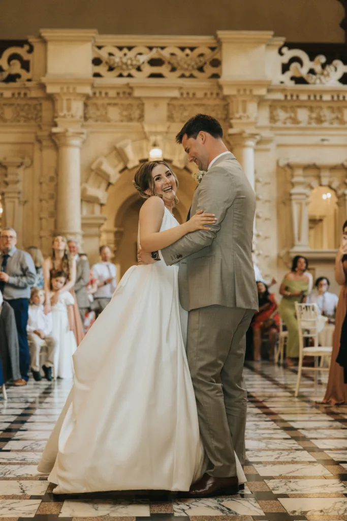 A bride in a white gown and groom in a grey suit share a joyful first dance at their Harlaxton Manor wedding. Guests in semi-formal attire watch from the ornate marble hall as the happy couple celebrate their love. © Aimee Lince Photography, Lincolnshire photographer