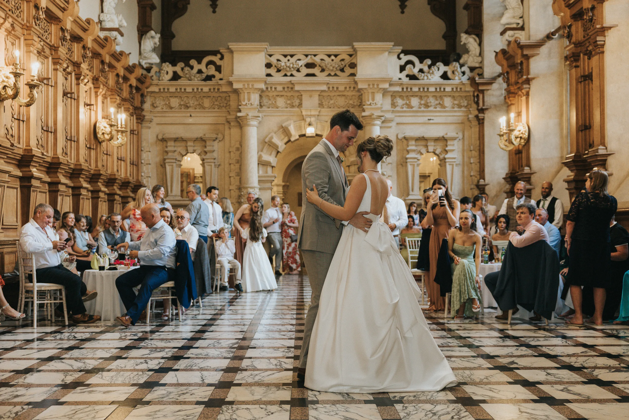 A bride and groom share their first dance in an ornate, high-ceilinged hall at a Harlaxton Manor Wedding, with marble floors and wood panelling. Guests watch and take photos as warm lighting and elegant decor create a festive, romantic atmosphere. © Aimee Lince Photography, Lincolnshire photographer