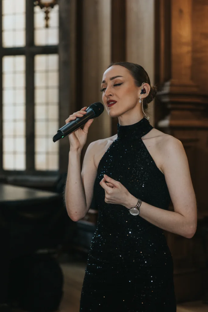 A woman in a sleeveless, black sequinned dress sings into a microphone with her eyes closed at a Harlaxton Manor wedding. She wears a watch and stud earrings, her brown hair slicked back, standing in an elegant, wood-panelled room with large windows. © Aimee Lince Photography, Lincolnshire photographer