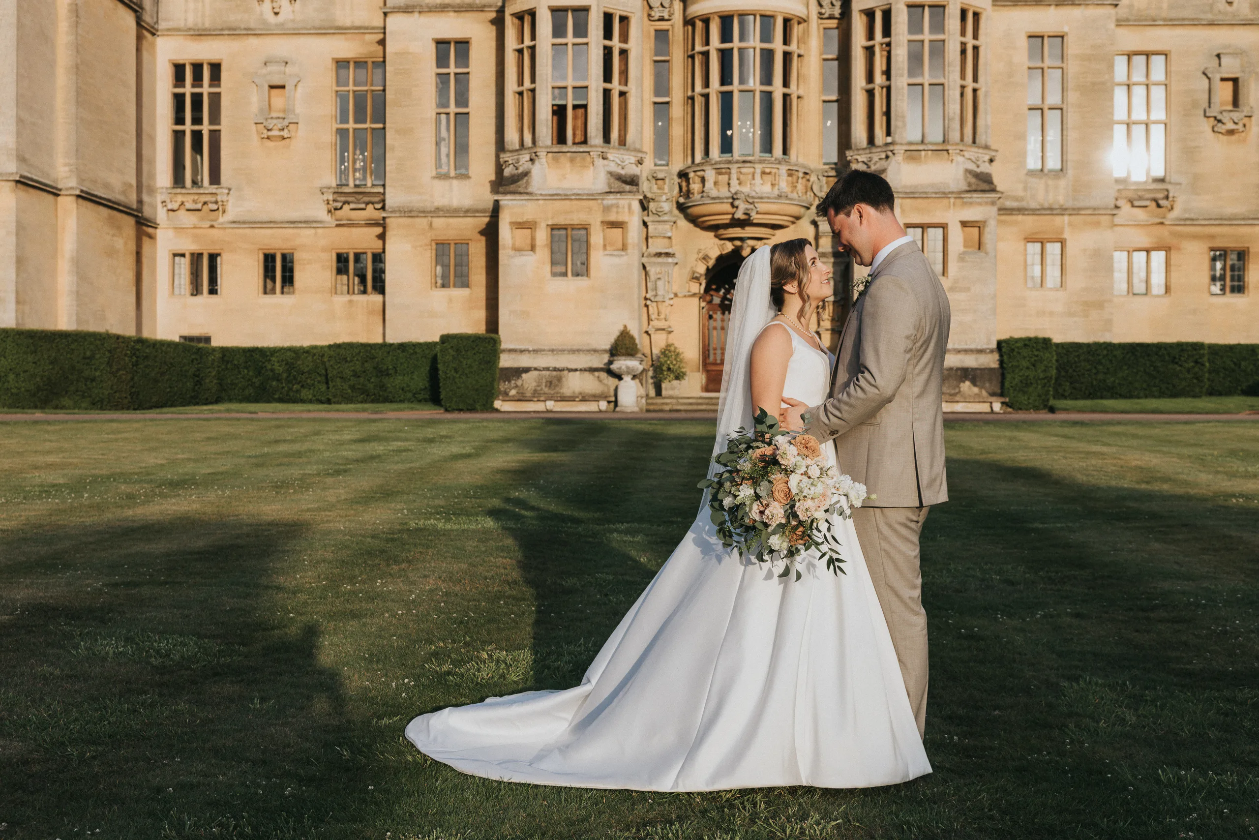 A bride in a white gown and veil stands facing a groom in a tan suit on a lawn at their Harlaxton Manor wedding, holding a bouquet of flowers. They touch foreheads as warm sunlight bathes the grand historic building behind them. © Aimee Lince Photography, Lincolnshire photographer