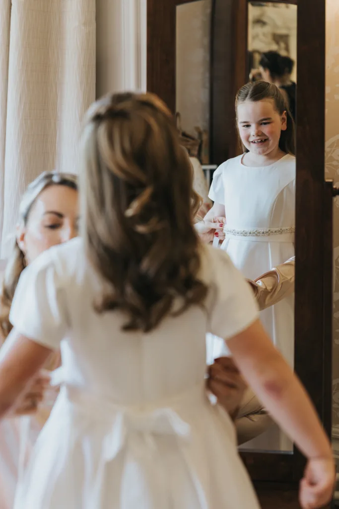 A young girl in a white dress smiles at her reflection in a tall mirror, her loose curls styled with care. An adult helps adjust her dress from behind, capturing a joyful, intimate moment before the Harlaxton Manor Wedding. © Aimee Lince Photography, Lincolnshire photographer
