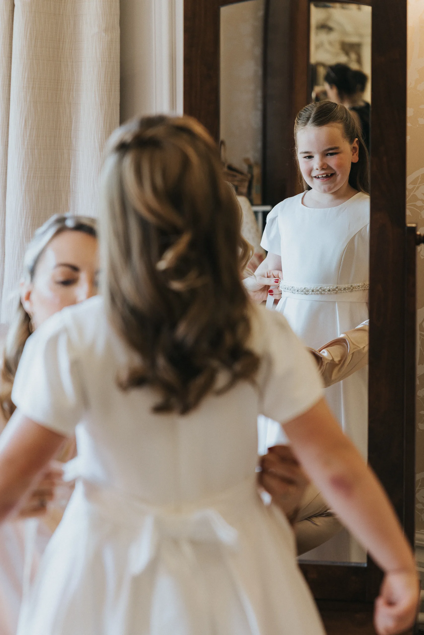 A young girl in a white dress smiles at her reflection in a tall mirror, her loose curls styled with care. An adult helps adjust her dress from behind, capturing a joyful, intimate moment before the Harlaxton Manor Wedding. © Aimee Lince Photography, Lincolnshire photographer